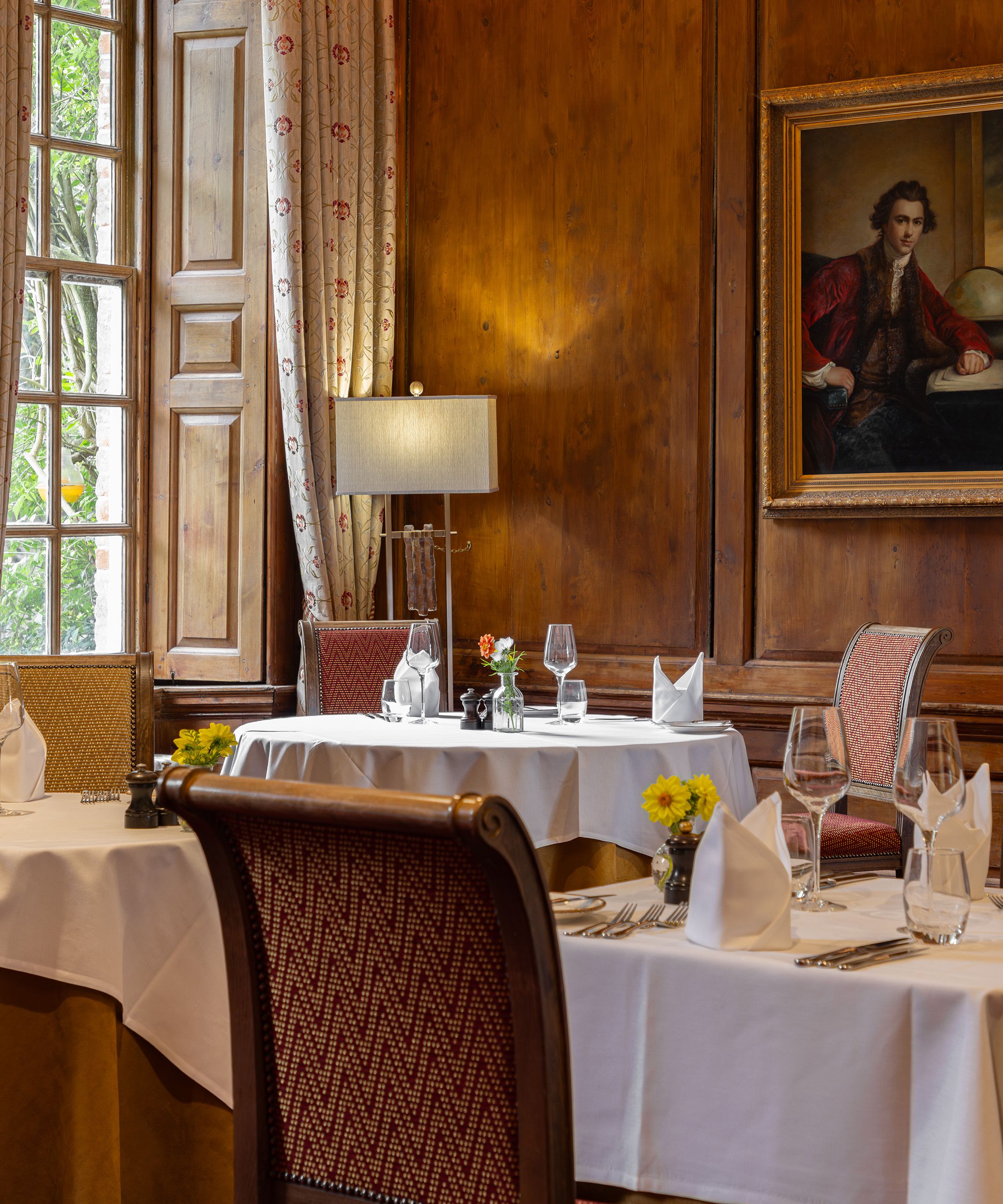 Elegant restaurant dining room with white tablecloths, ornate chairs, and classic paintings on wooden-paneled walls.