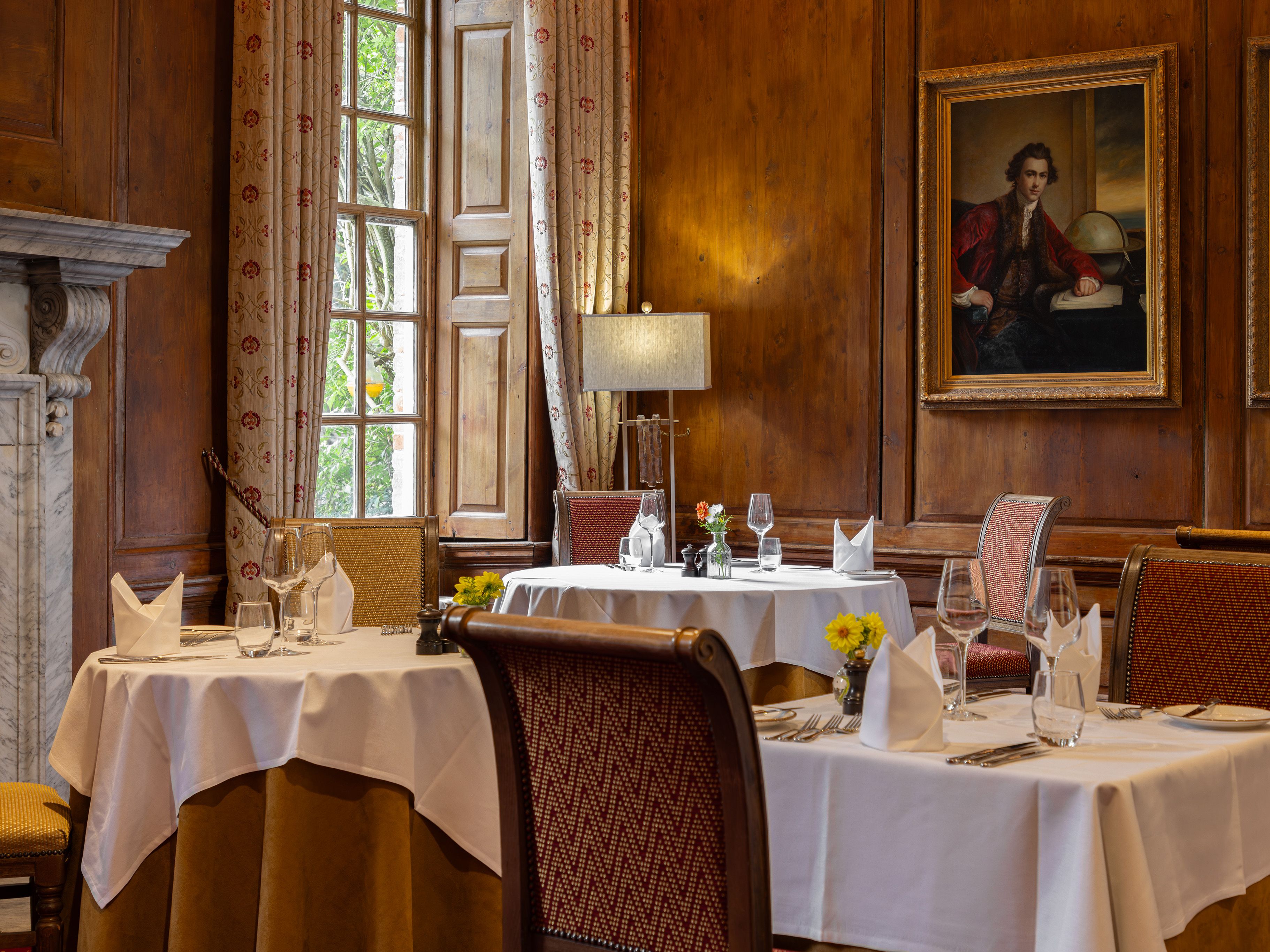 Elegant restaurant dining room with white tablecloths, ornate chairs, and classic paintings on wooden-paneled walls.