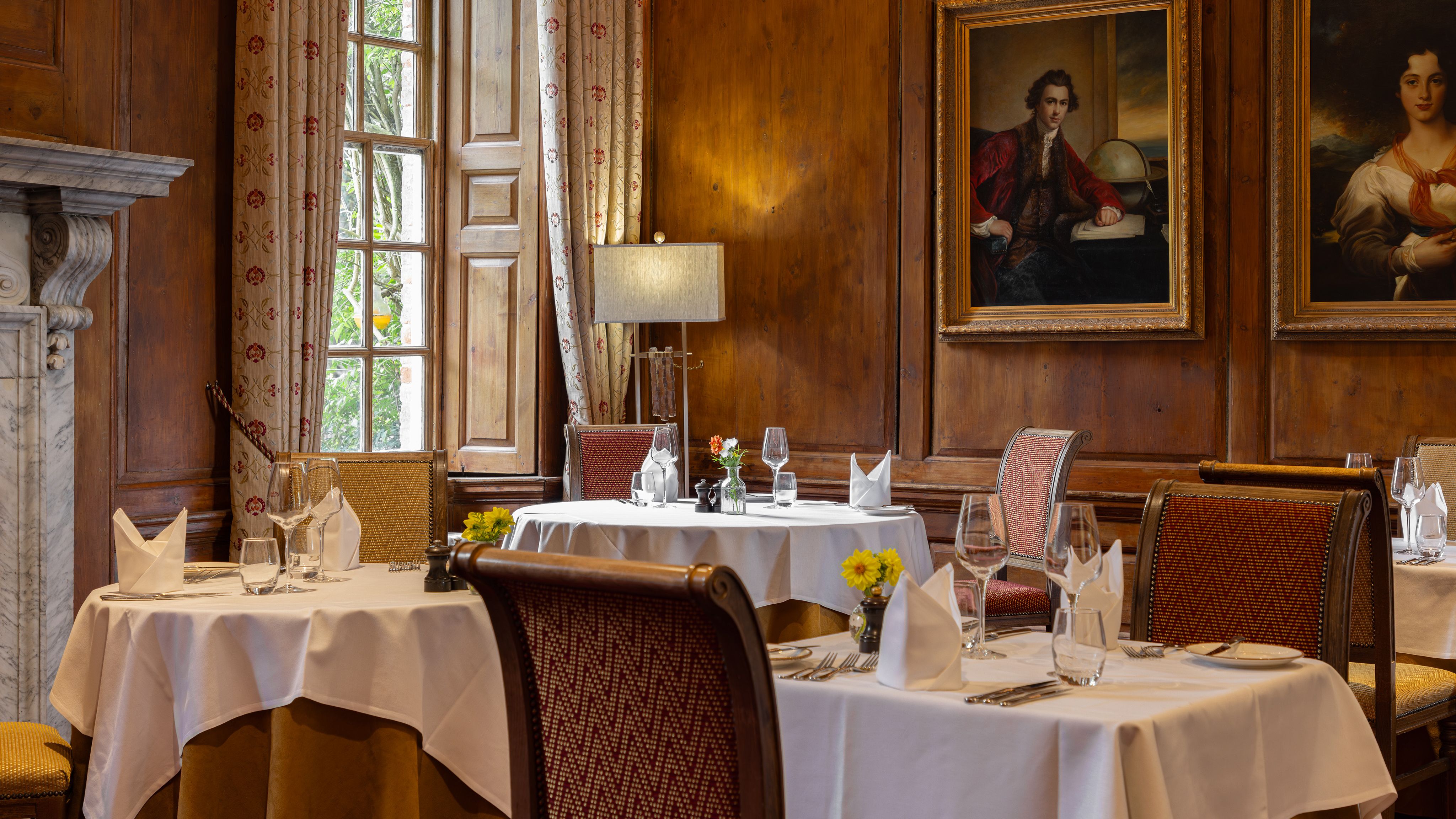 Elegant restaurant dining room with white tablecloths, ornate chairs, and classic paintings on wooden-paneled walls.