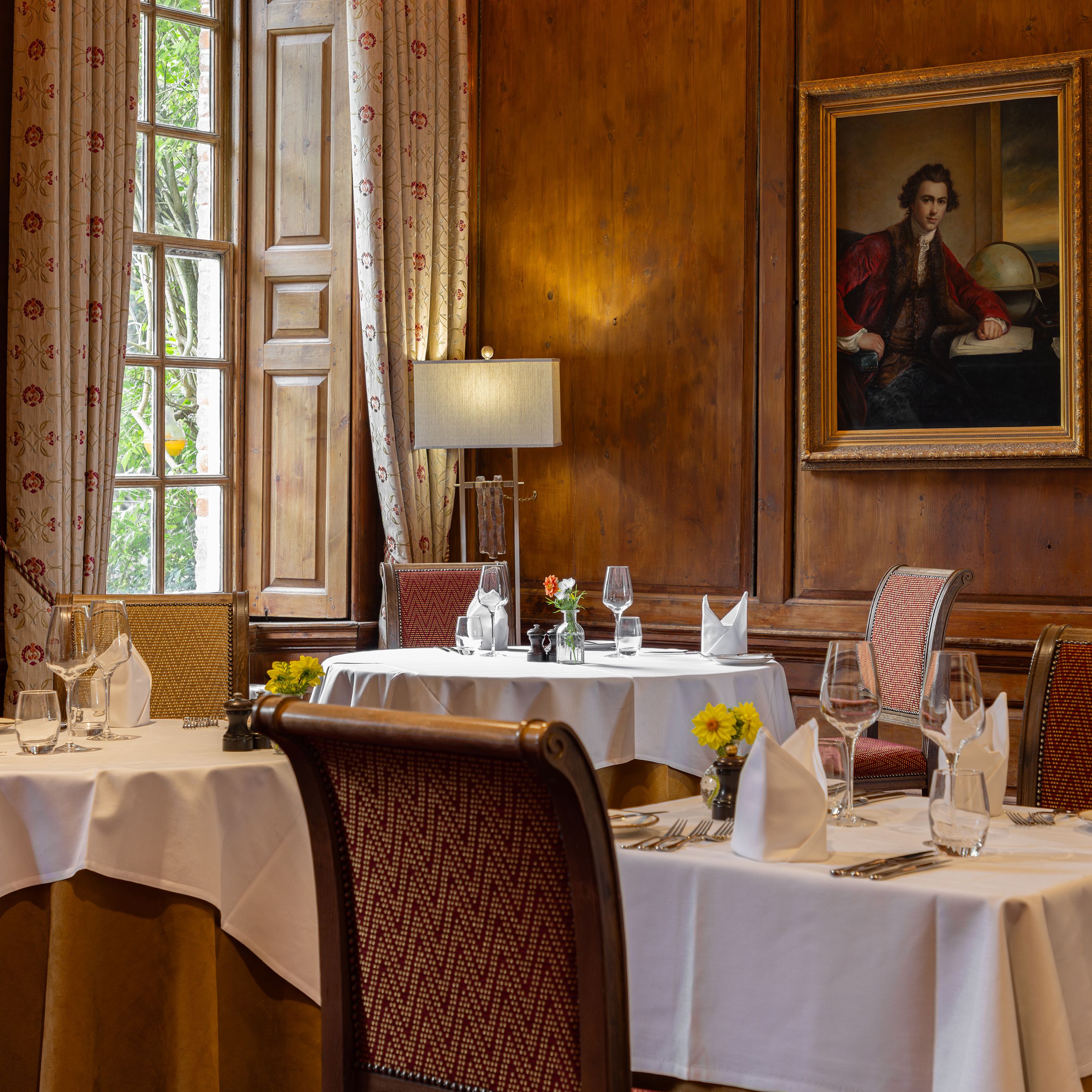 Elegant restaurant dining room with white tablecloths, ornate chairs, and classic paintings on wooden-paneled walls.