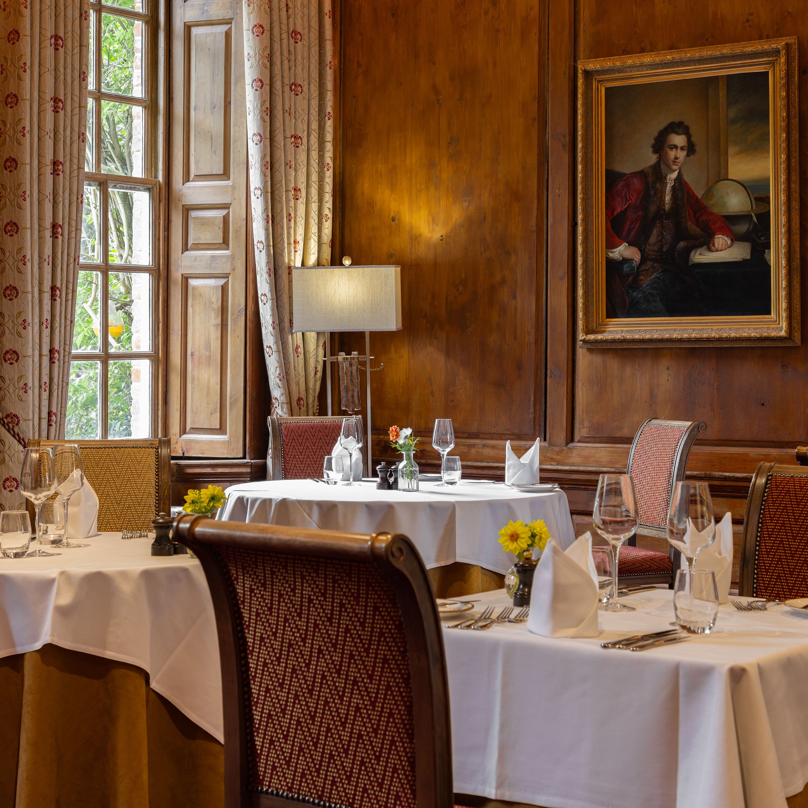 Elegant restaurant dining room with white tablecloths, ornate chairs, and classic paintings on wooden-paneled walls.