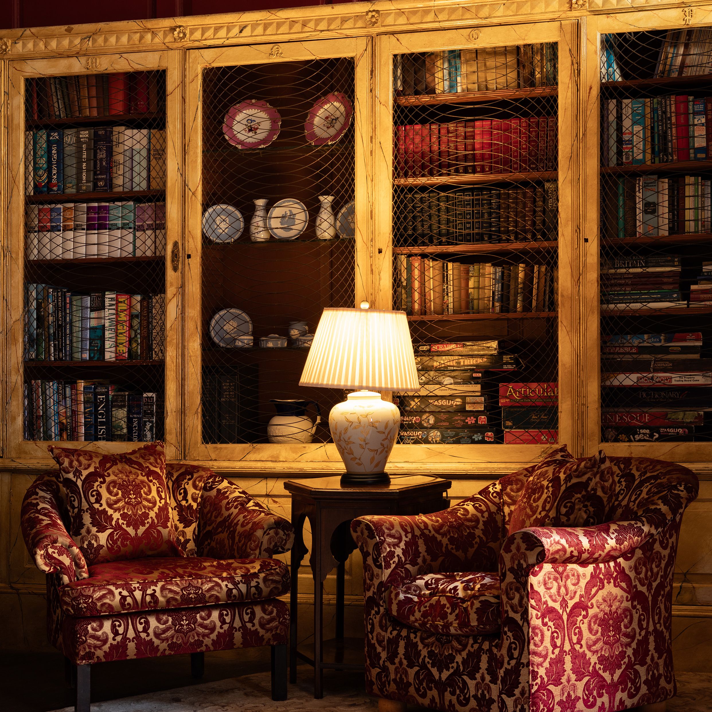 Cozy reading nook with two red armchairs, a lamp, and a bookshelf filled with books and ceramics.
