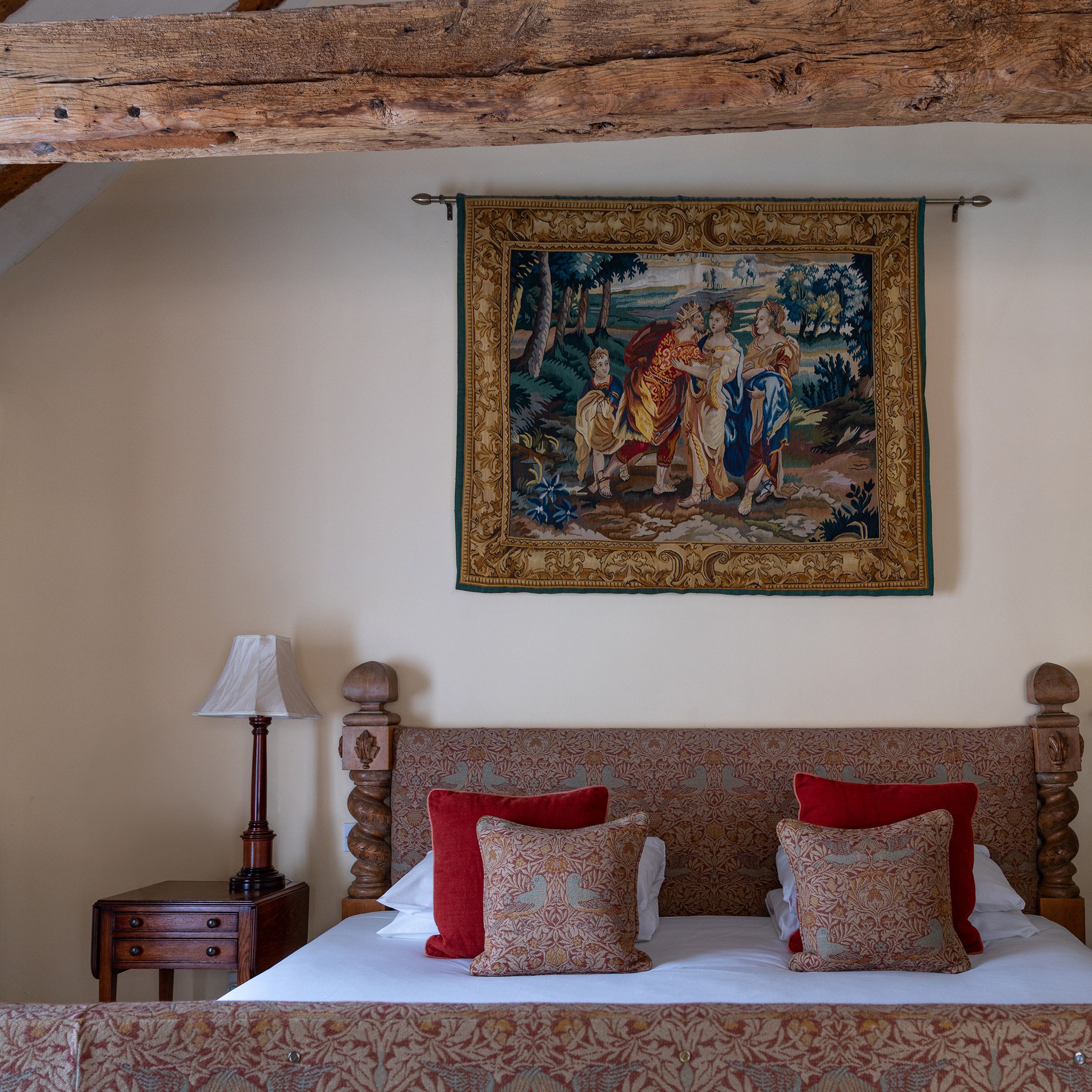 A cozy bedroom with a wooden bed, patterned pillows, a side table with a lamp, and a decorative tapestry hanging above the bed.