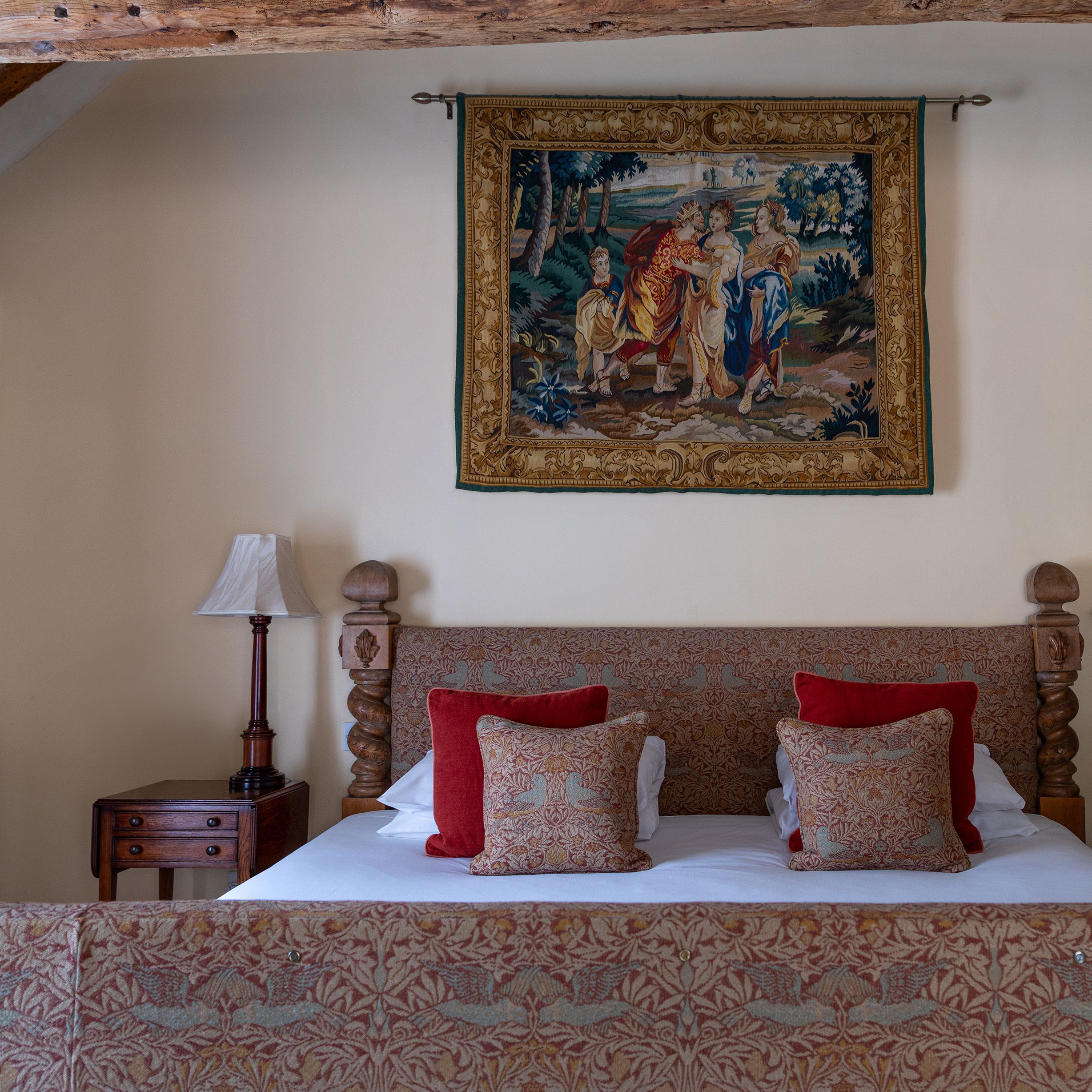 A cozy bedroom with a wooden bed, patterned pillows, a side table with a lamp, and a decorative tapestry hanging above the bed.