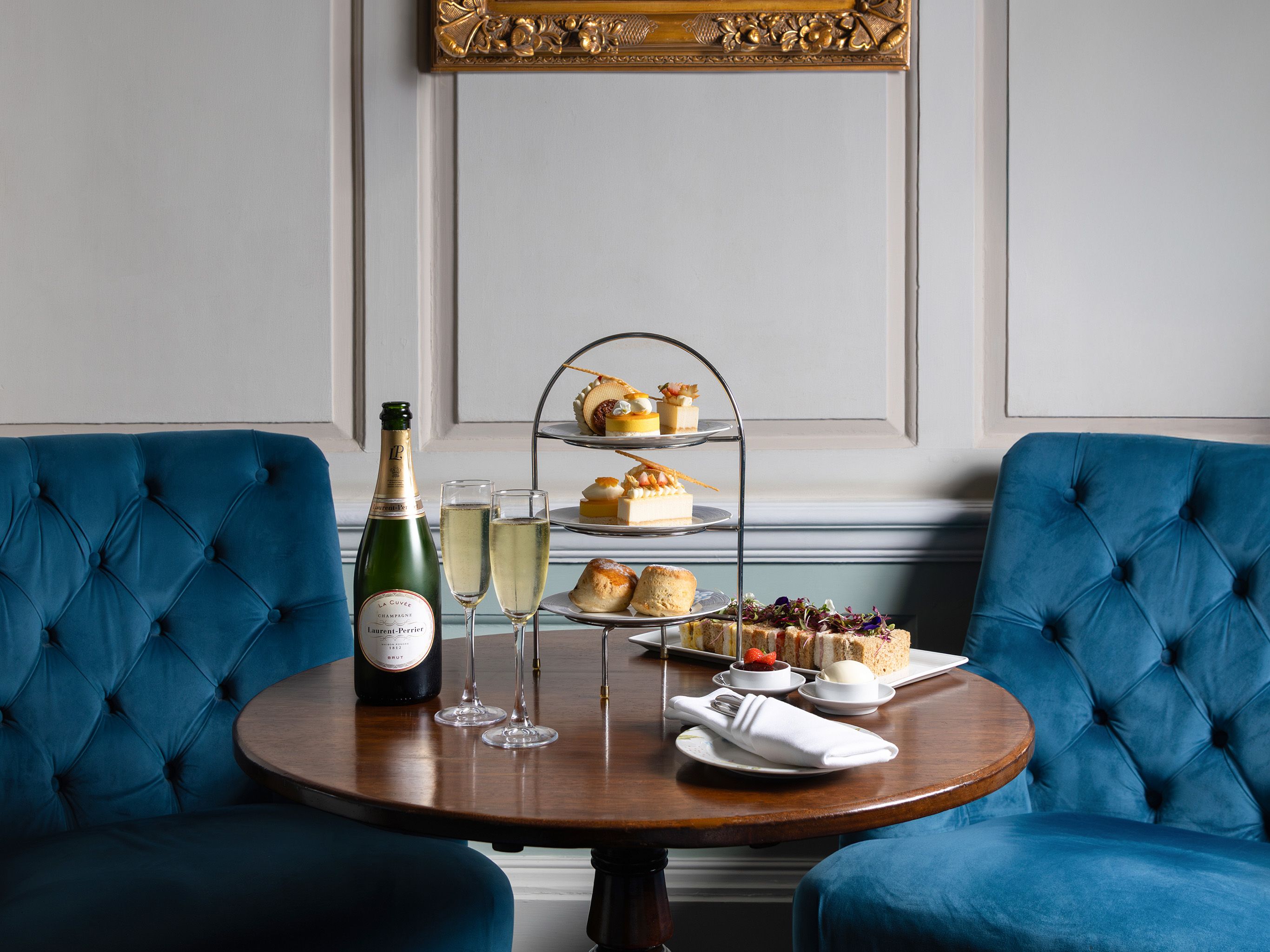 Elegant afternoon tea setup with champagne, pastries, and scones on a wooden table between two blue velvet chairs, under a framed portrait painting.