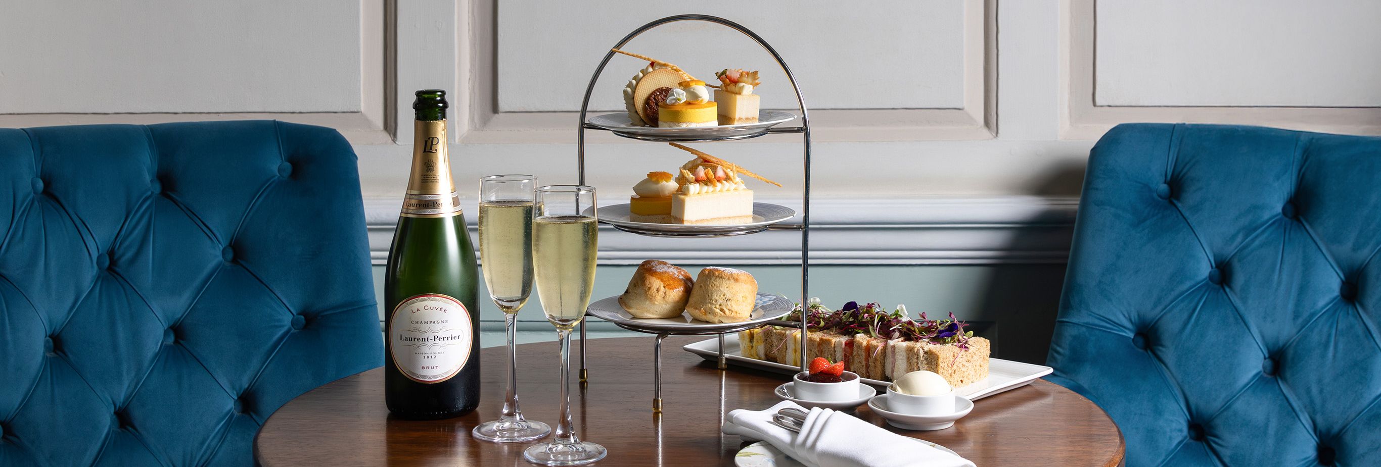 Elegant afternoon tea setup with champagne, pastries, and scones on a wooden table between two blue velvet chairs, under a framed portrait painting.