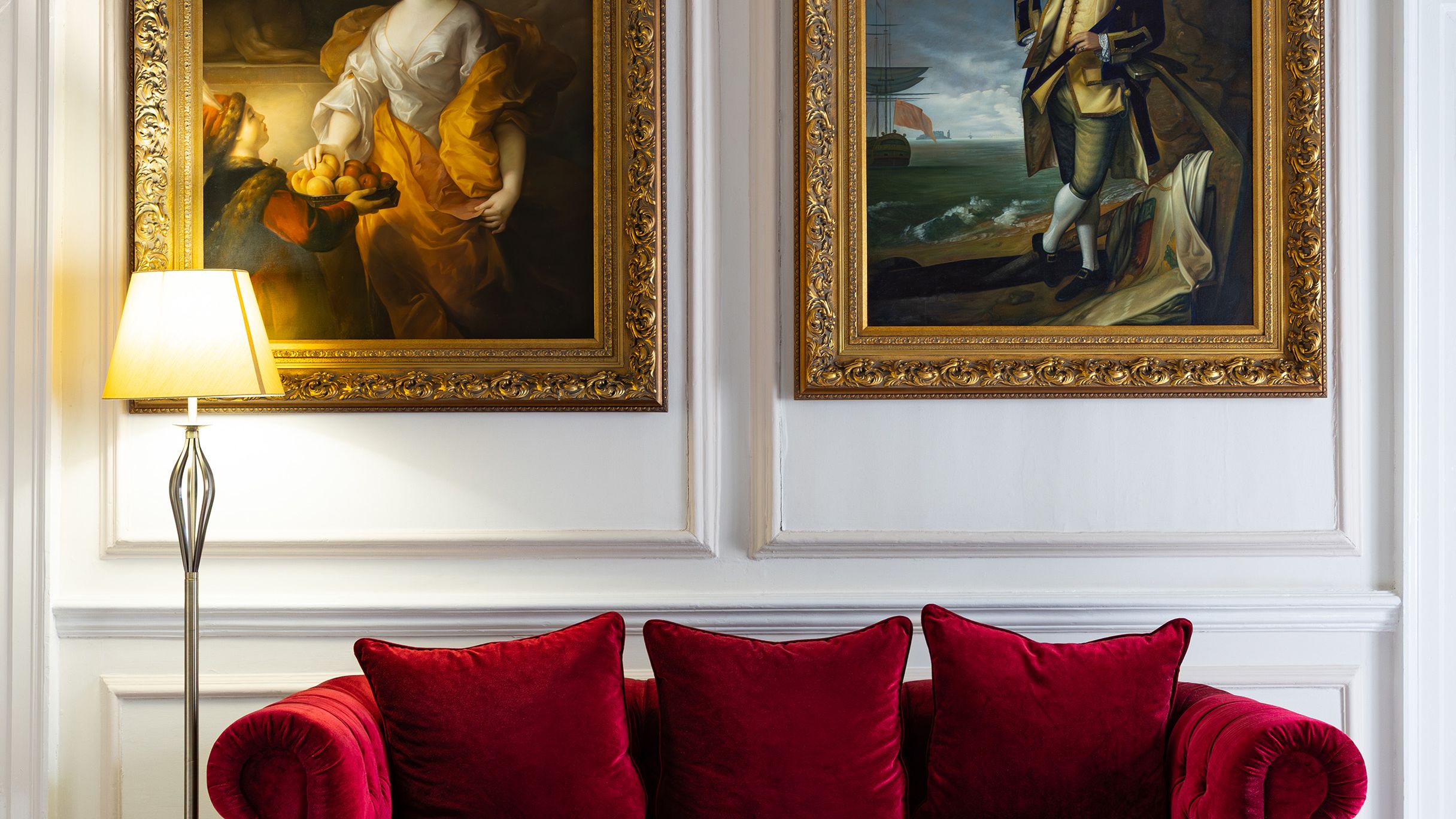 Elegant living room with a red velvet sofa, two classical gold-framed portraits, and a floor lamp