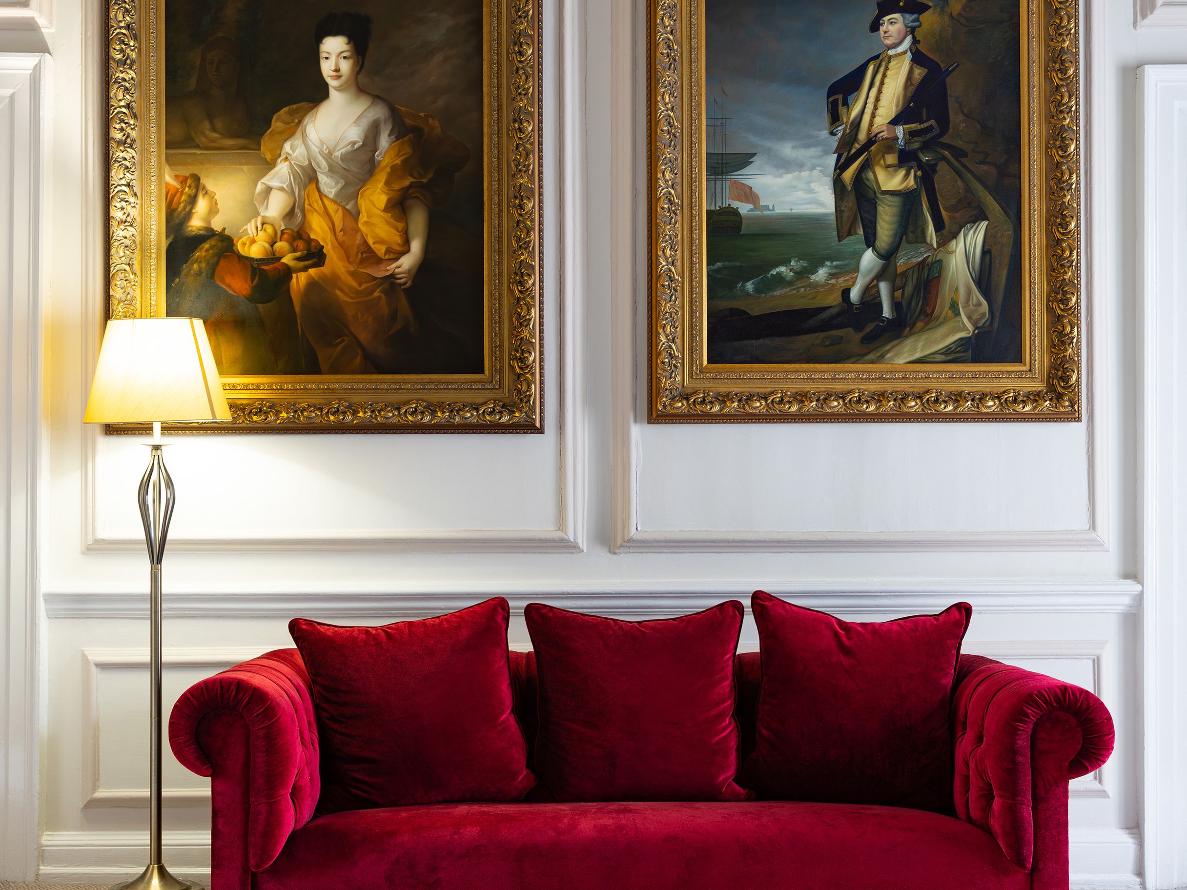 Elegant living room with a red velvet sofa, two classical gold-framed portraits, and a floor lamp
