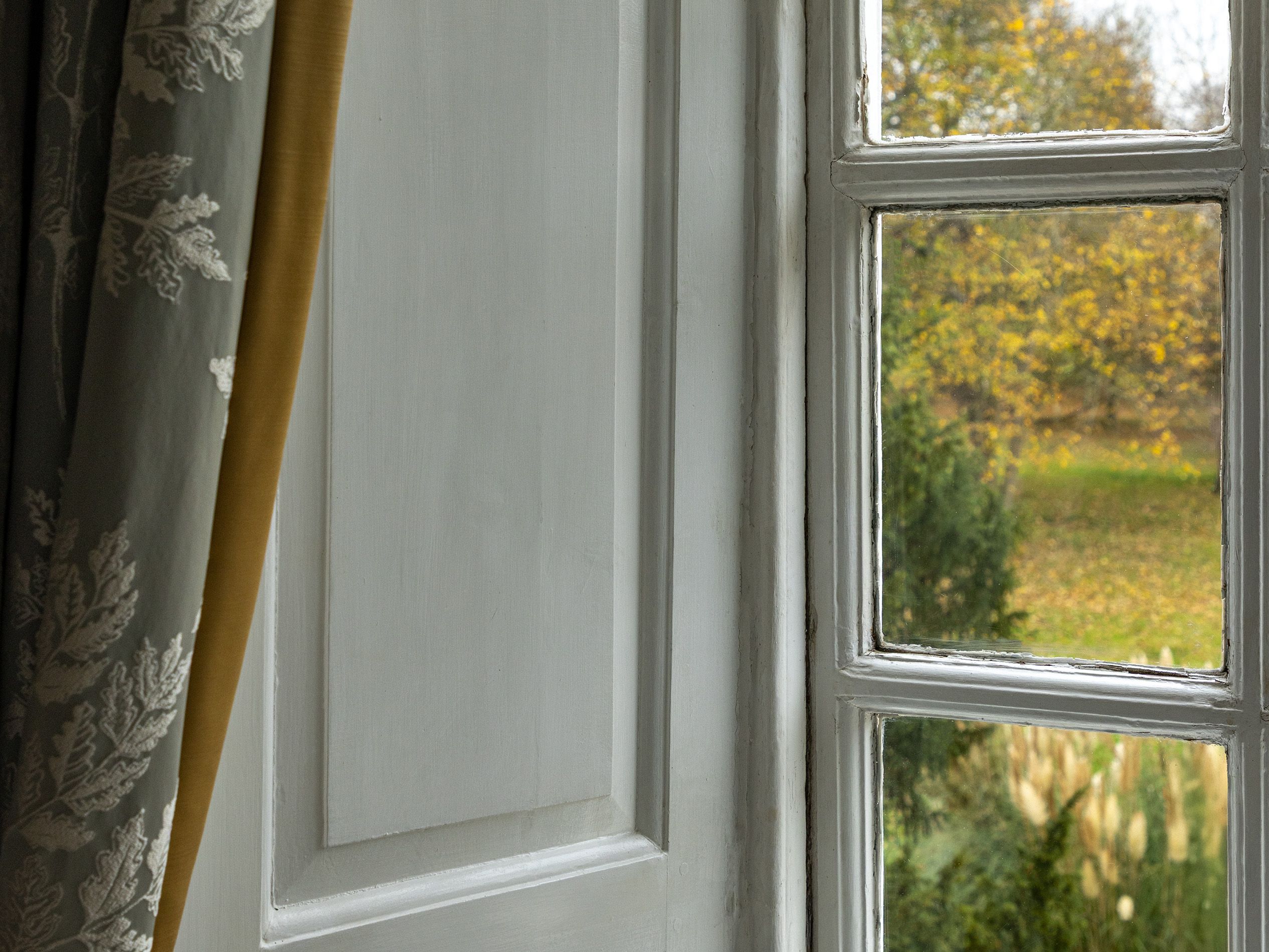 Close-up of a window with white wooden frames and partially drawn floral curtains, overlooking a garden with trees.