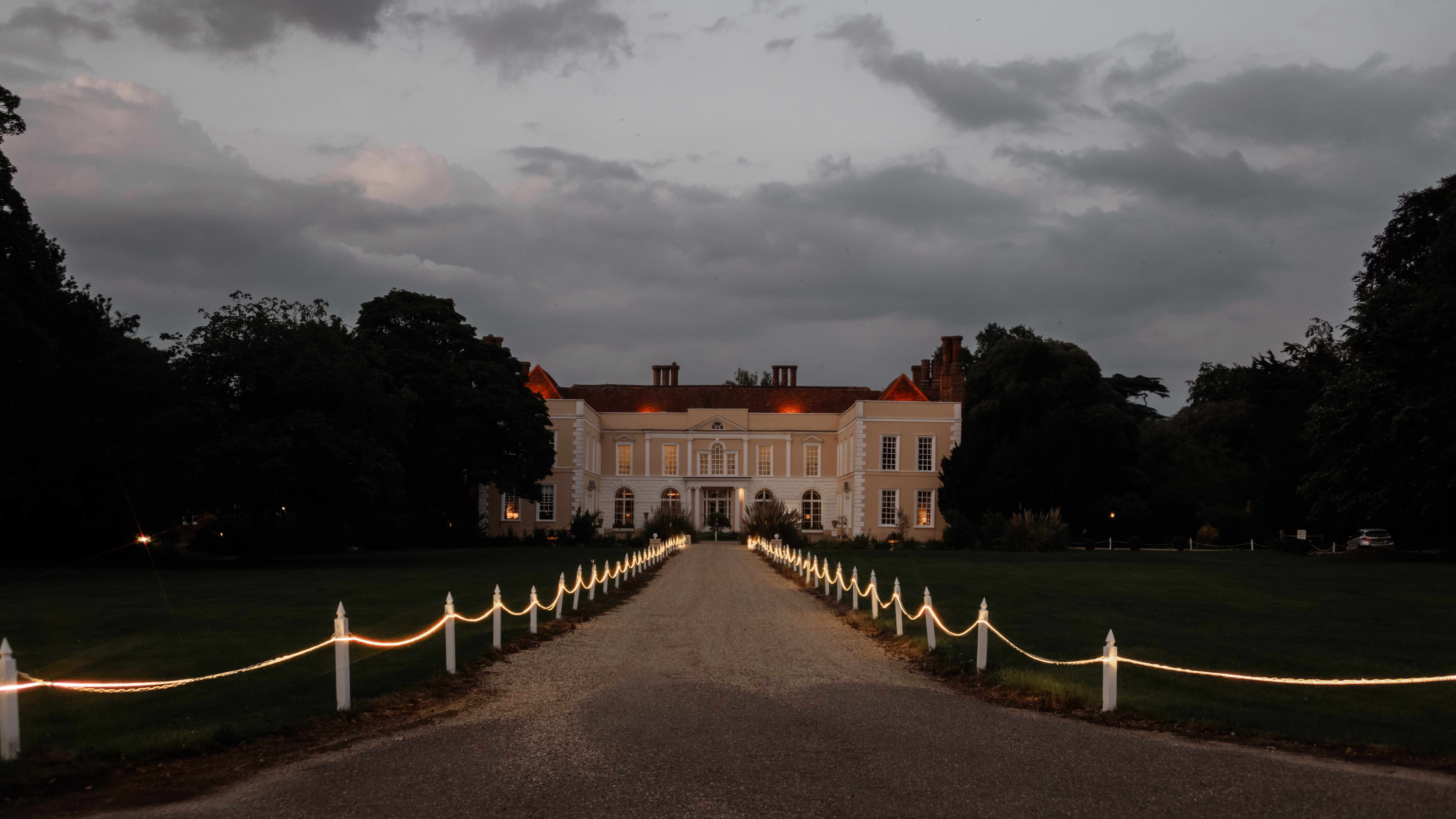 Large manor house at dusk with a driveway lined with illuminated rope lights.