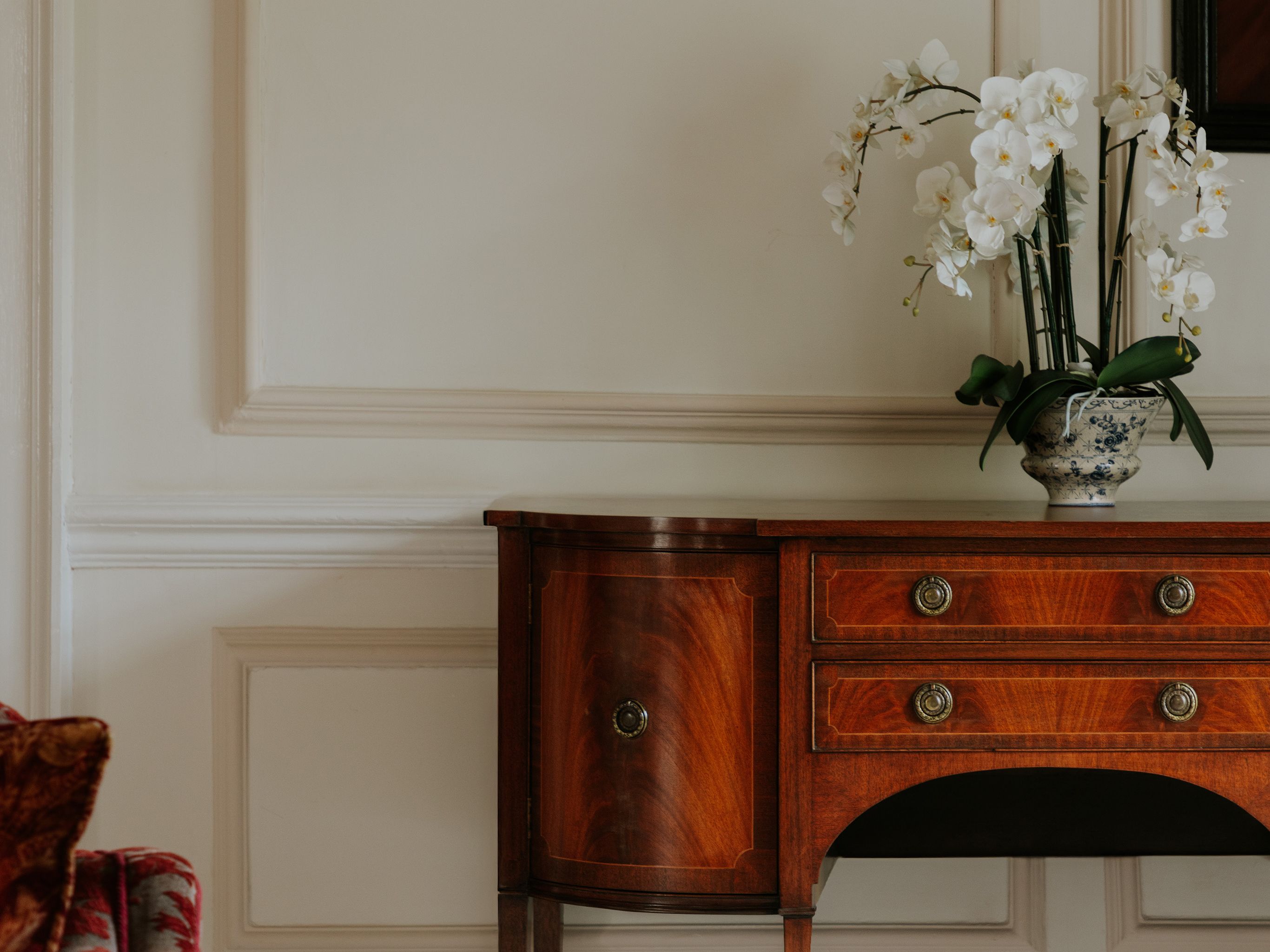 Elegant wooden sideboard with brass handles and vase of white orchids against a cream paneled wall.