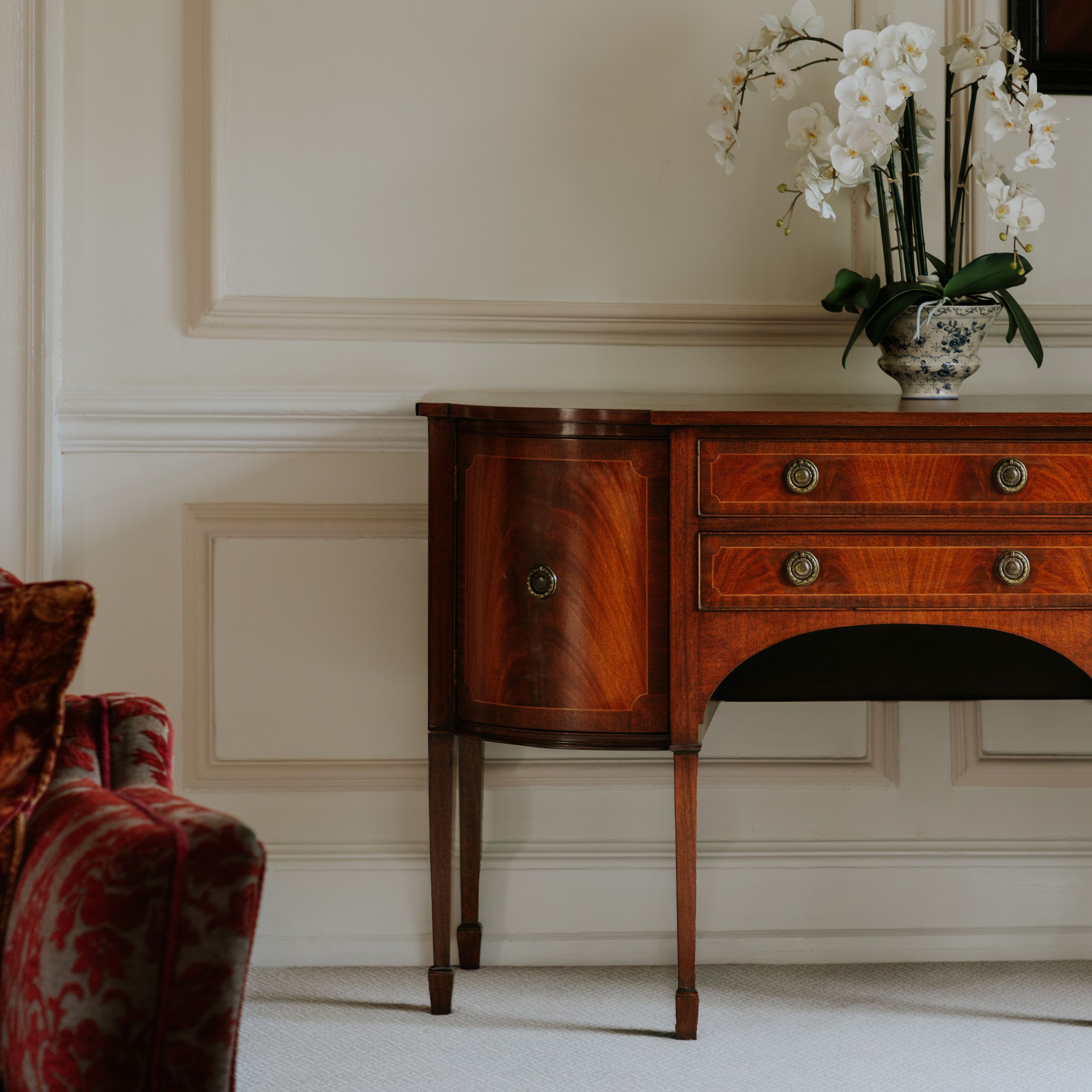 Elegant wooden sideboard with brass handles and vase of white orchids against a cream paneled wall.