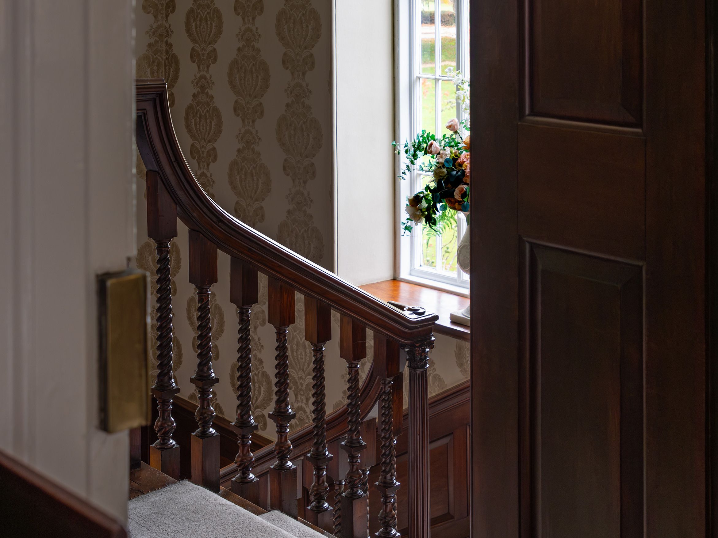 Interior view of a staircase with dark wooden bannister and ornate wallpaper, next to a large arched window with a flower arrangement on the sill.