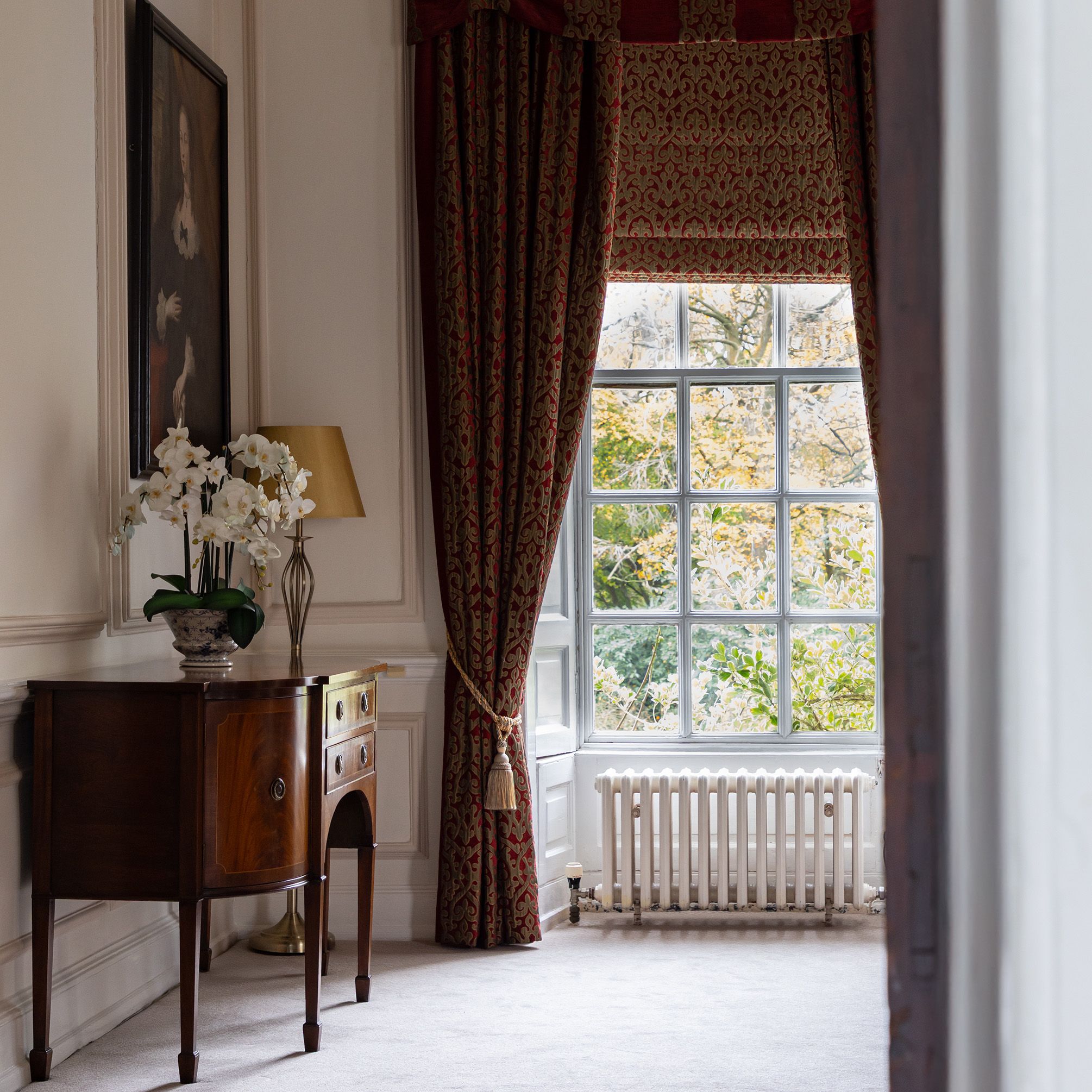 Elegant room with a large window, red curtains, a wooden side table, and a floral arrangement.