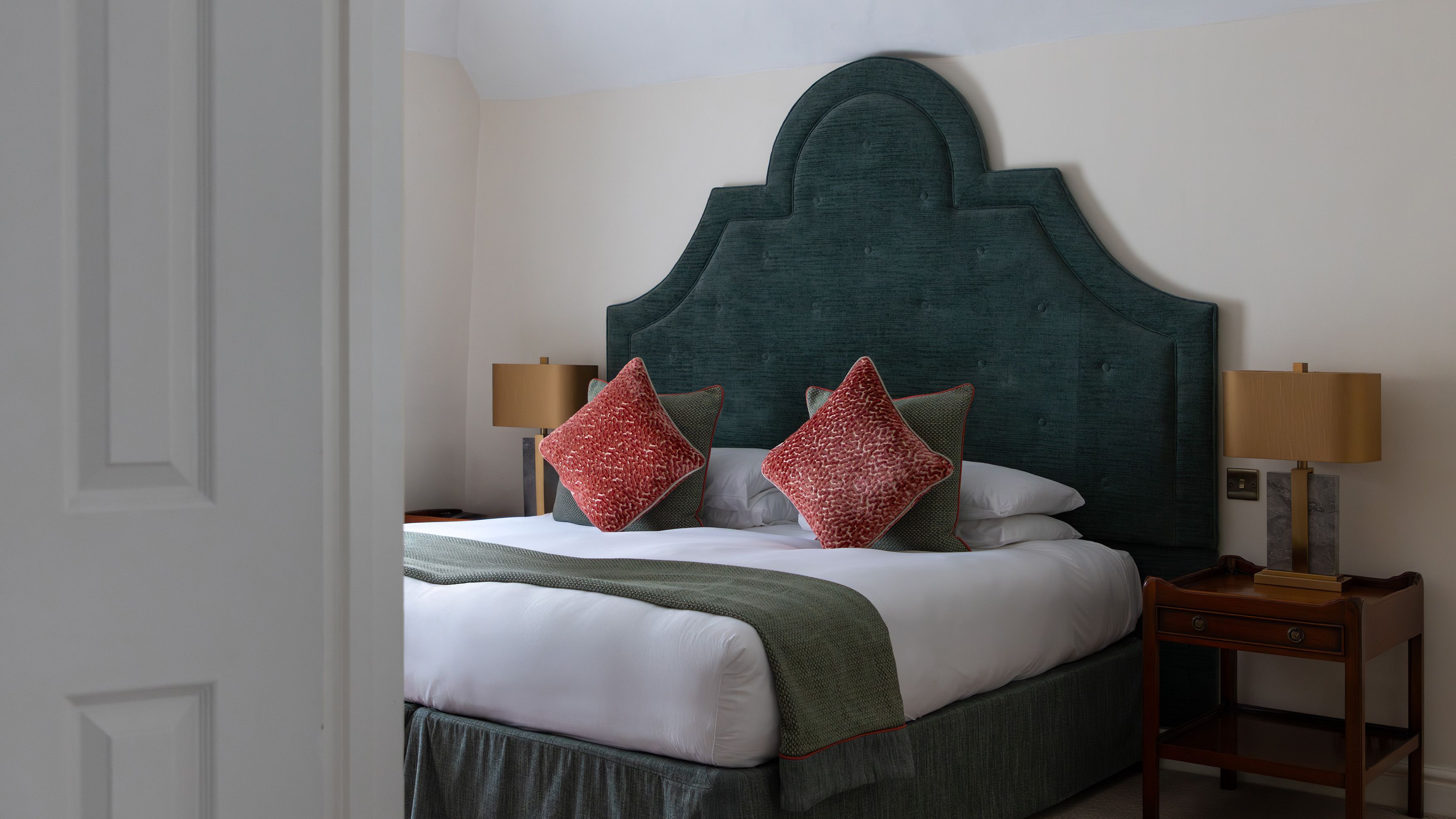 A neatly made bed with a large green headboard, red throw pillows, and side tables with lamps.
