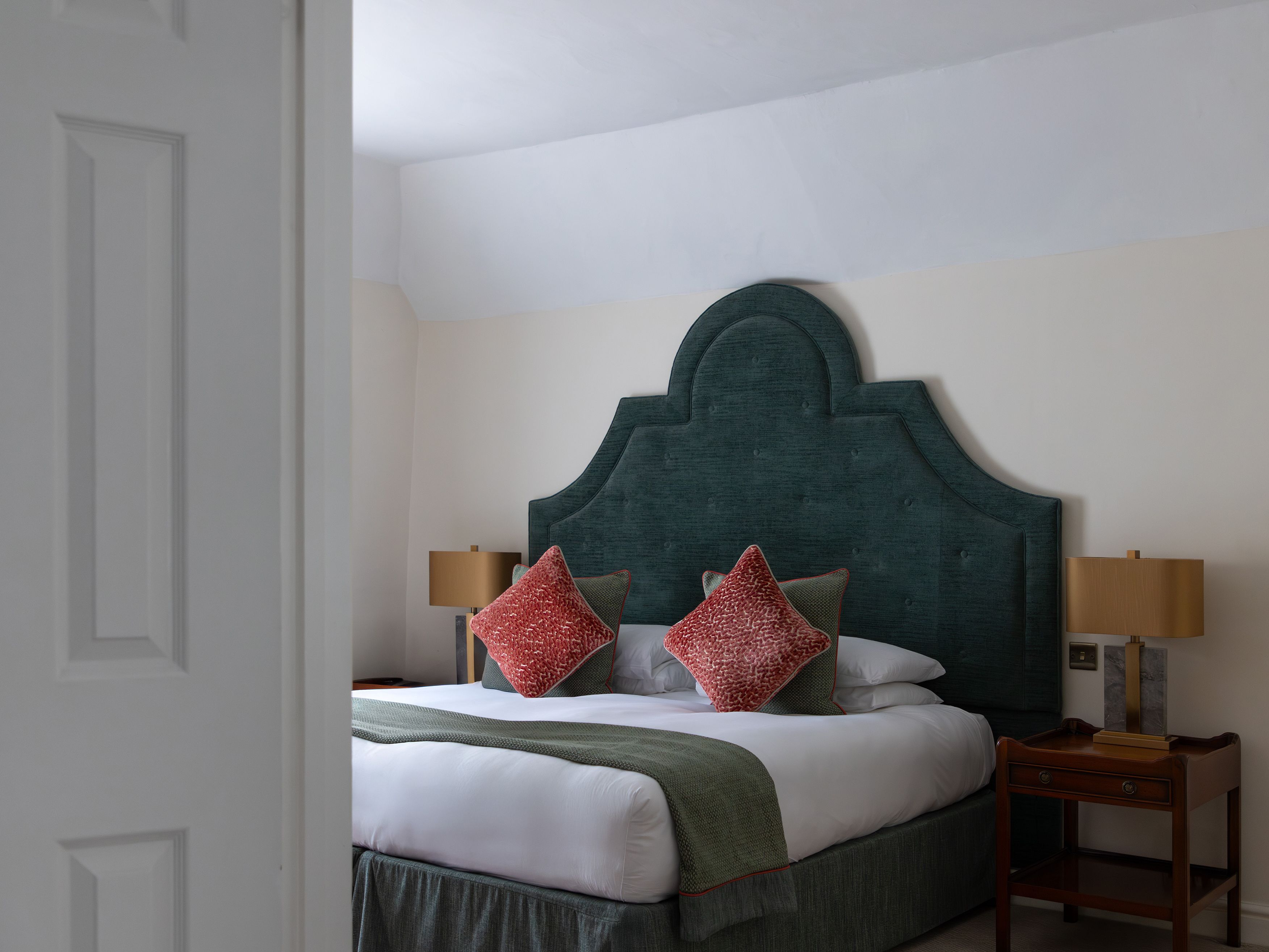 A neatly made bed with a large green headboard, red throw pillows, and side tables with lamps.