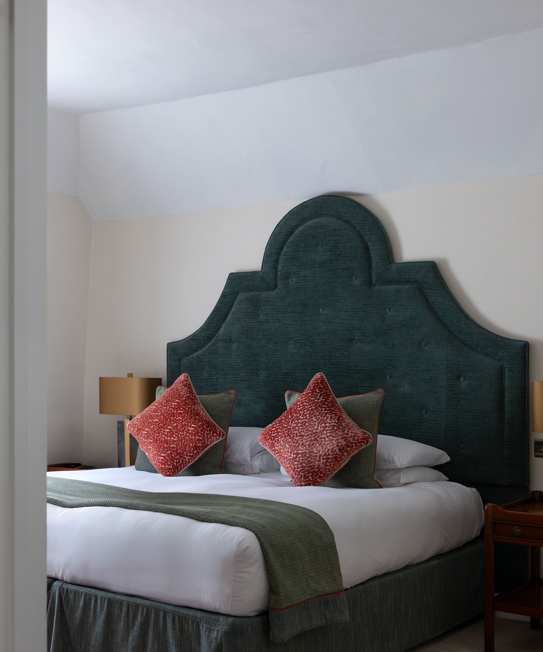 A neatly made bed with a large green headboard, red throw pillows, and side tables with lamps.