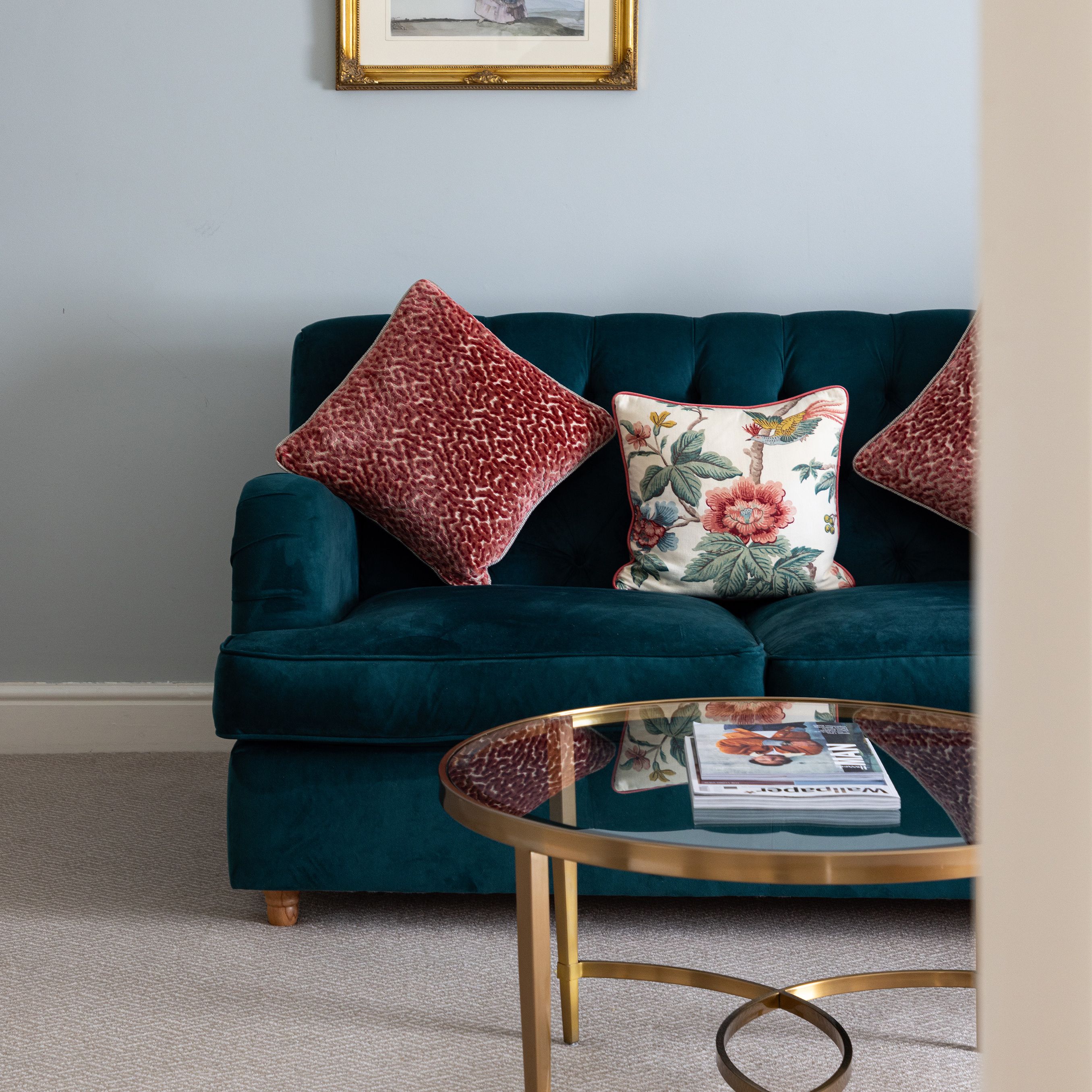 A stylish living room with a teal sofa, decorative pillows, a gold-framed artwork, and a round glass coffee table.