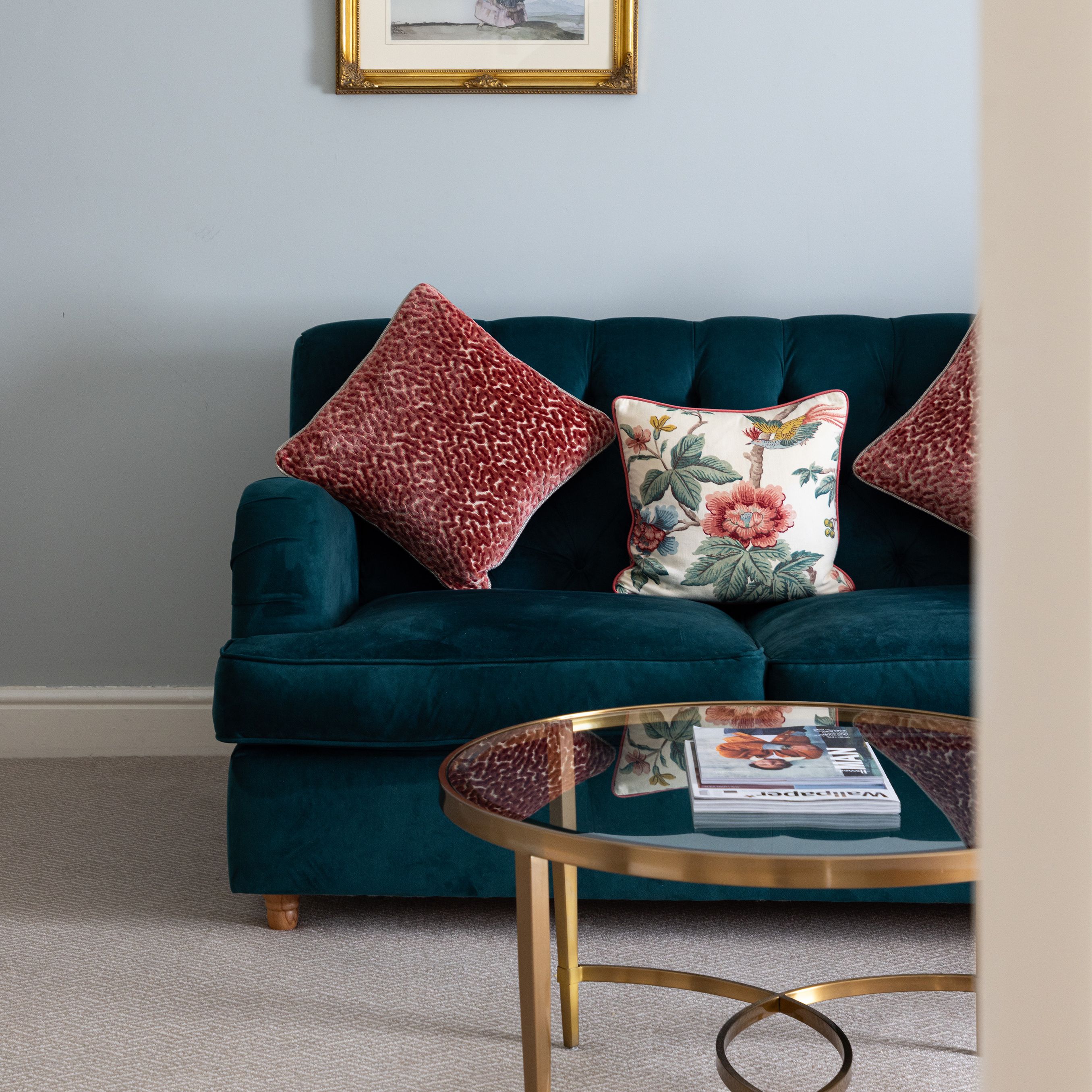 A stylish living room with a teal sofa, decorative pillows, a gold-framed artwork, and a round glass coffee table.