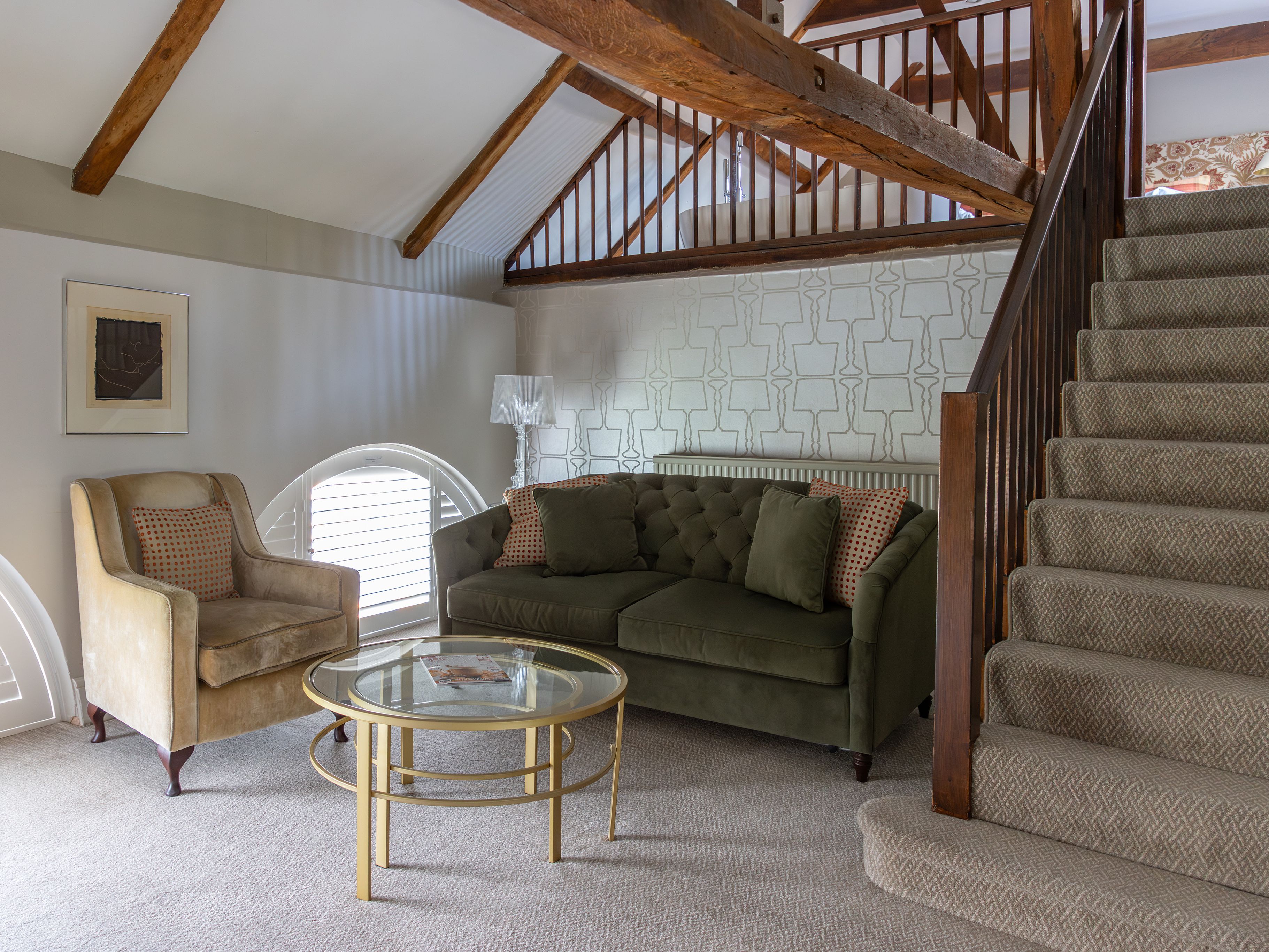 A cozy living room with a green sofa, beige armchair, glass coffee table, and carpeted staircase leading to a loft area.