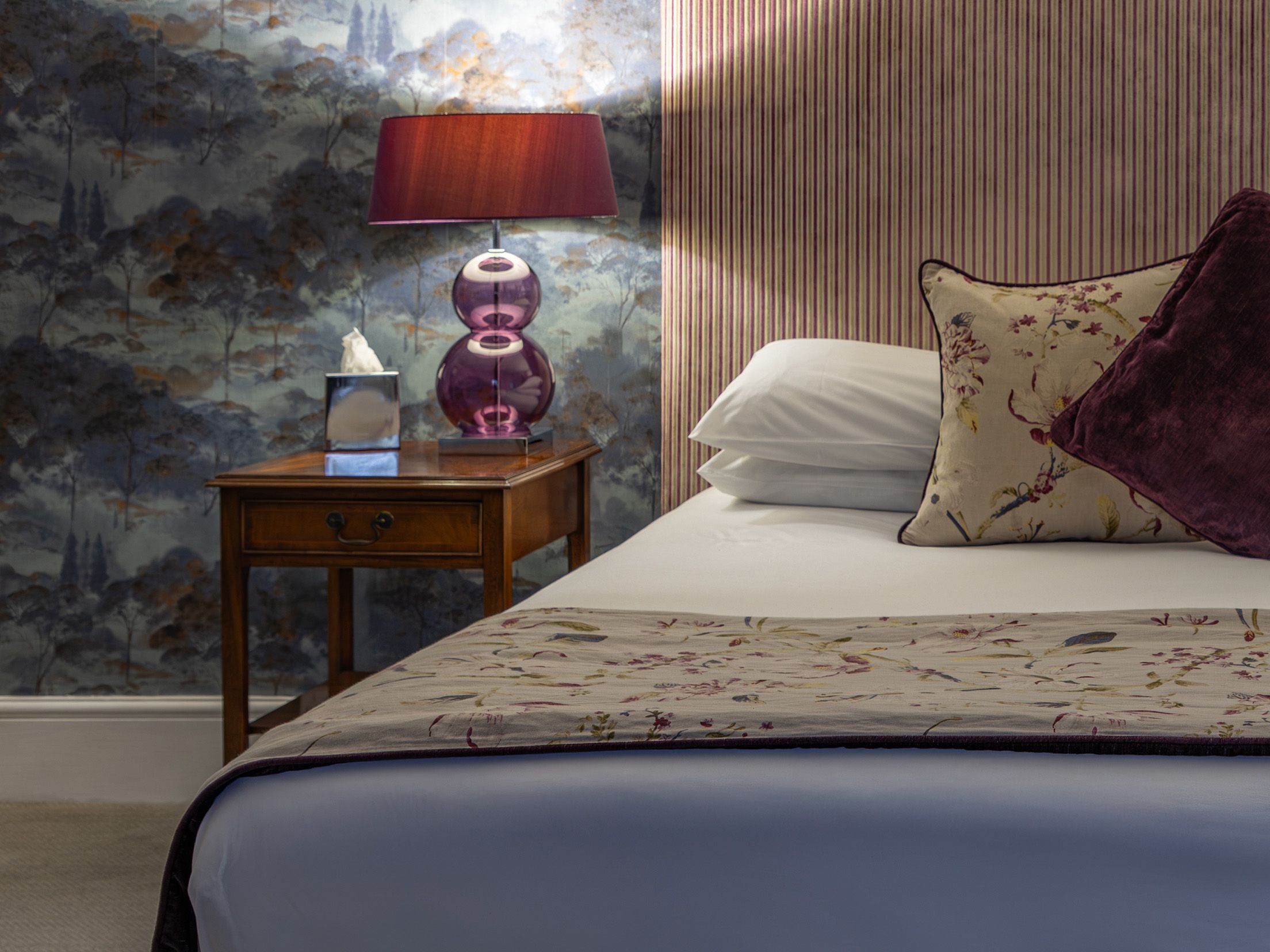 Cozy bedroom with patterned wallpaper, a bed with decorative pillows, and a bedside table with a lamp.