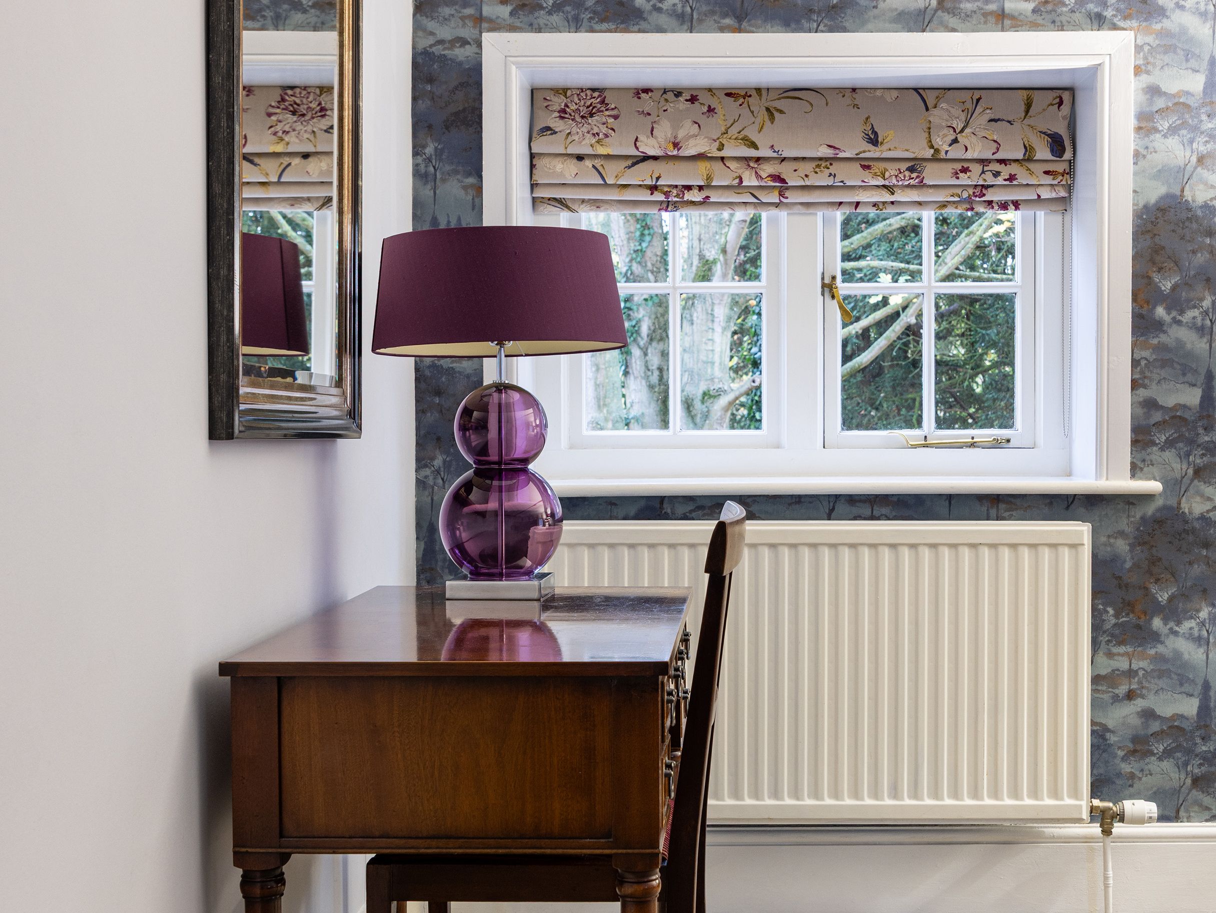 Elegant writing desk with a purple lamp in front of a window with floral curtains and blue patterned wallpaper.