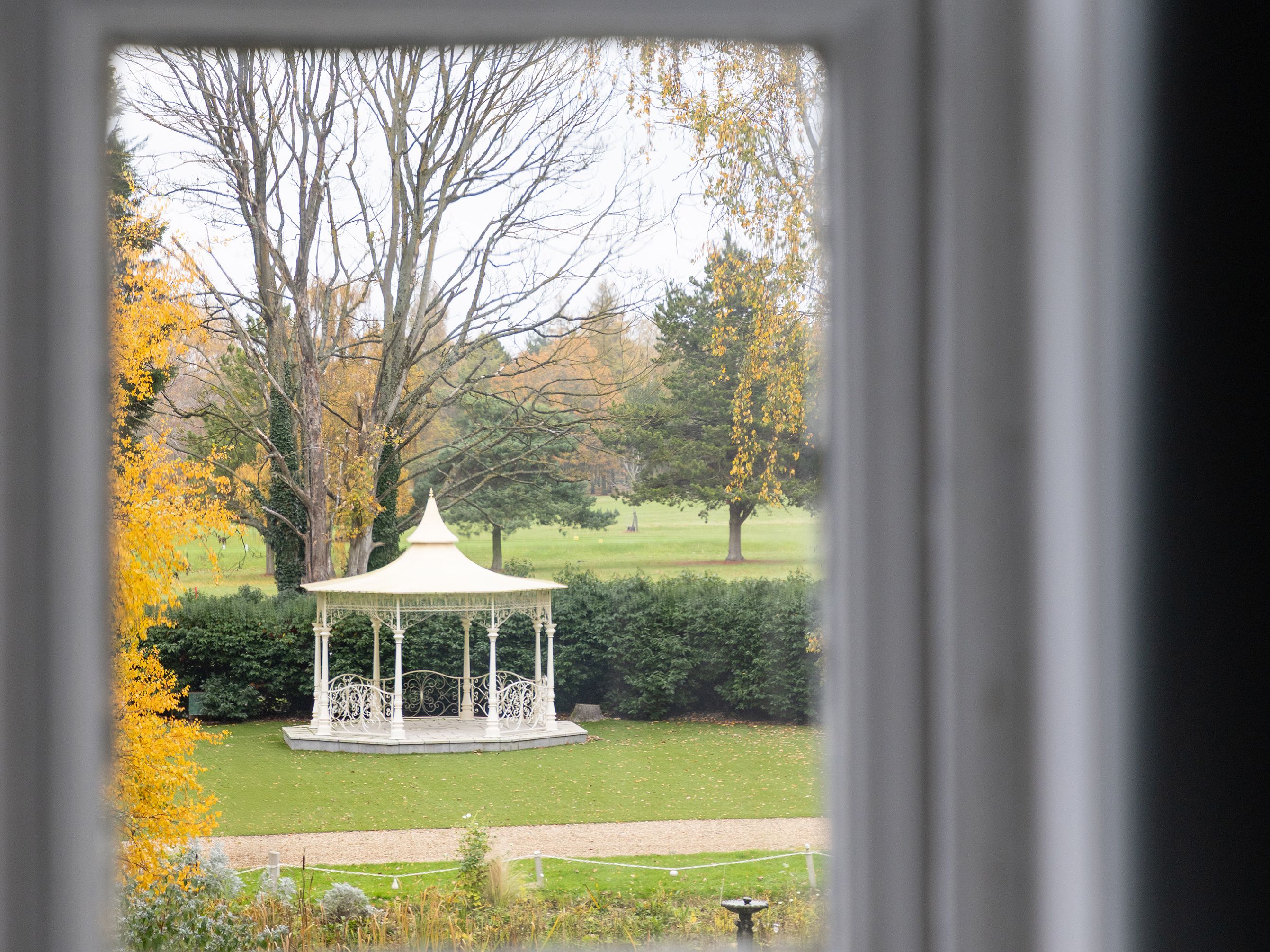 View through a window of a white gazebo in a garden surrounded by trees
