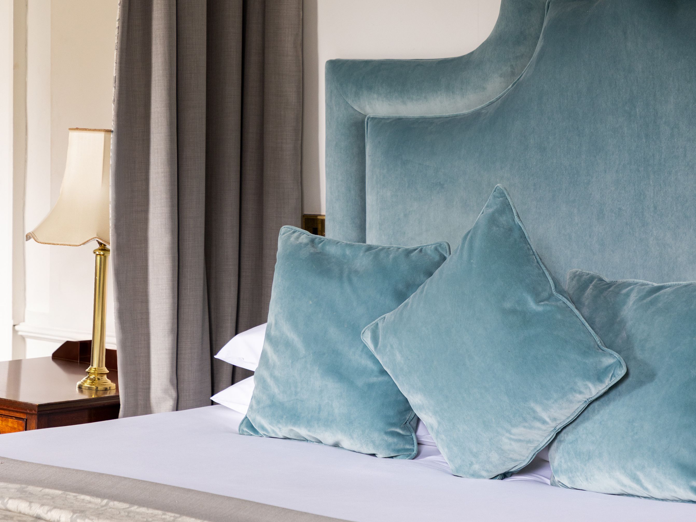 Elegant bedroom with a blue velvet upholstered headboard, matching pillows, gray canopy curtains, and a bedside lamp.