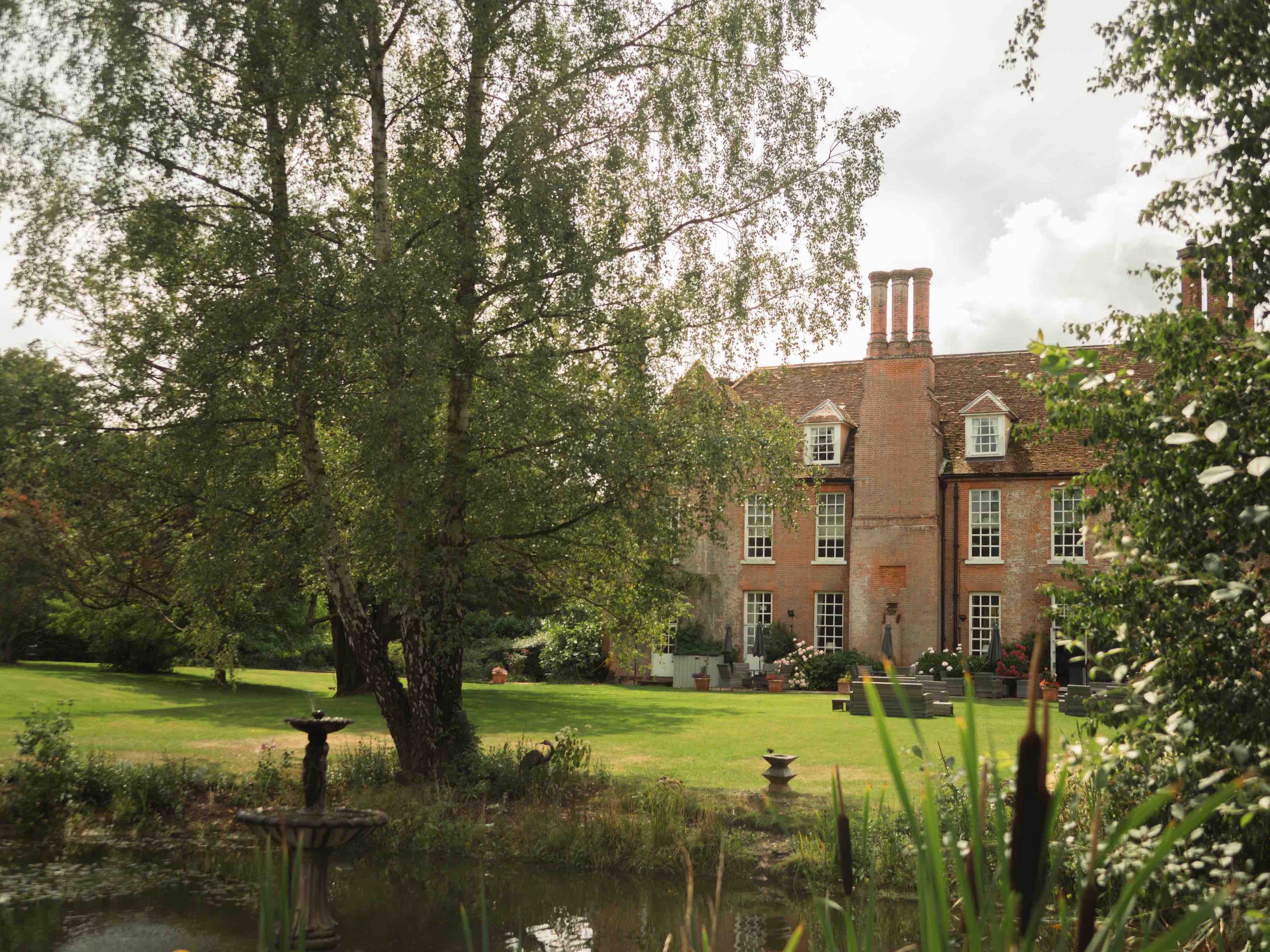 Large brick house with multiple chimneys, surrounded by a lush garden and pond