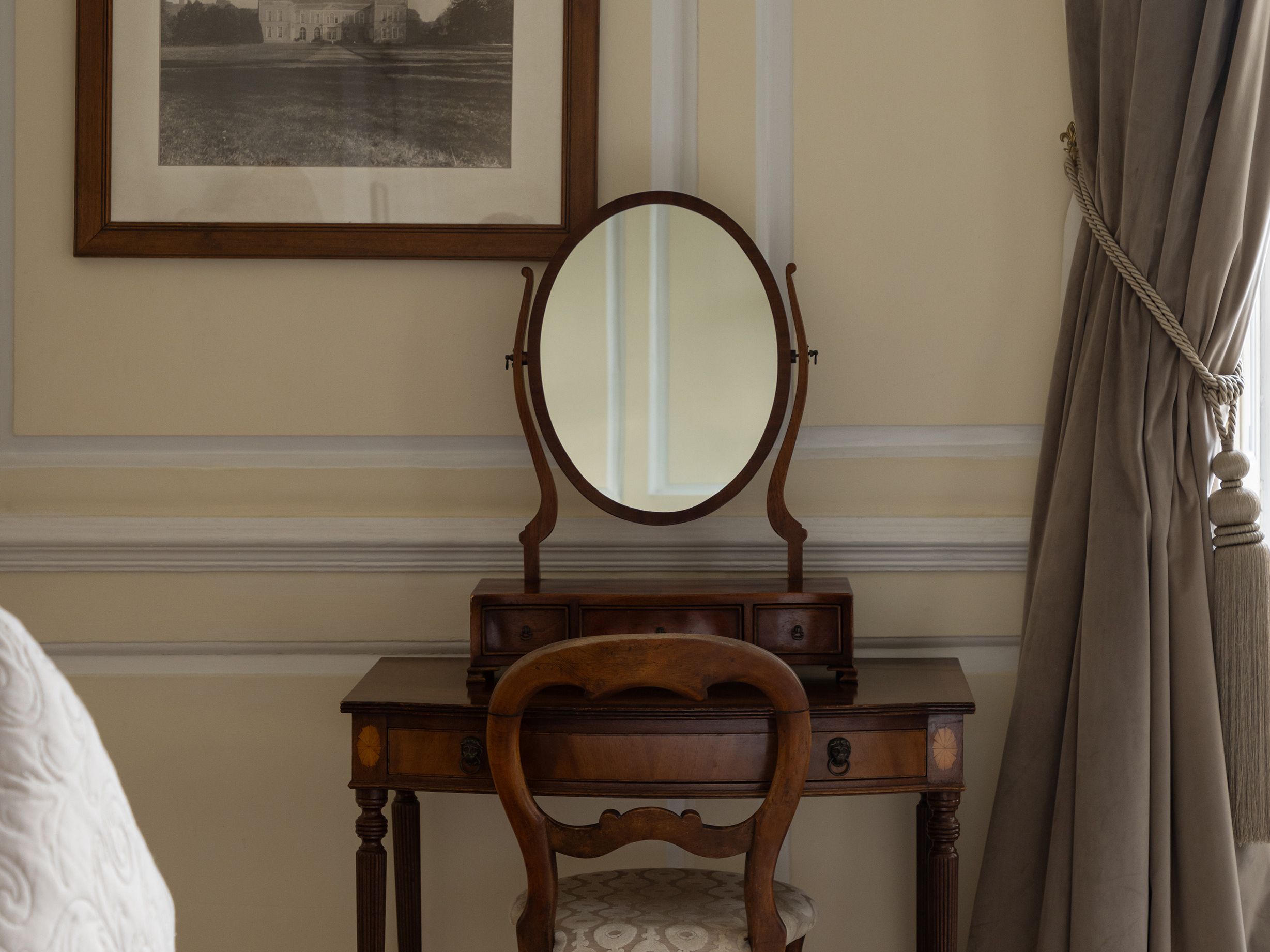 Antique wooden vanity with an oval mirror and chair against a wall, with a framed black and white photograph above and draped curtains to the side.