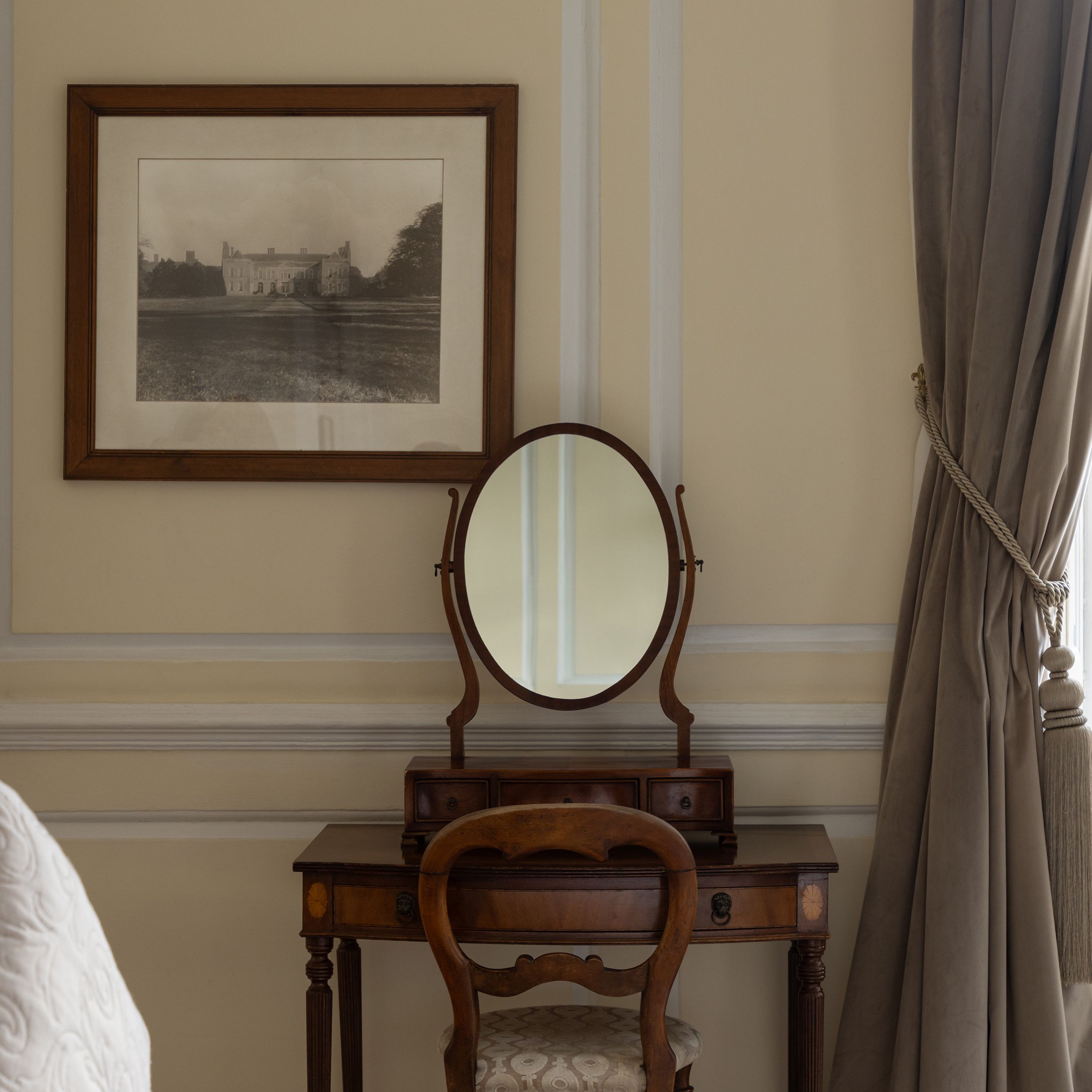 Antique wooden vanity with an oval mirror and chair against a wall, with a framed black and white photograph above and draped curtains to the side.