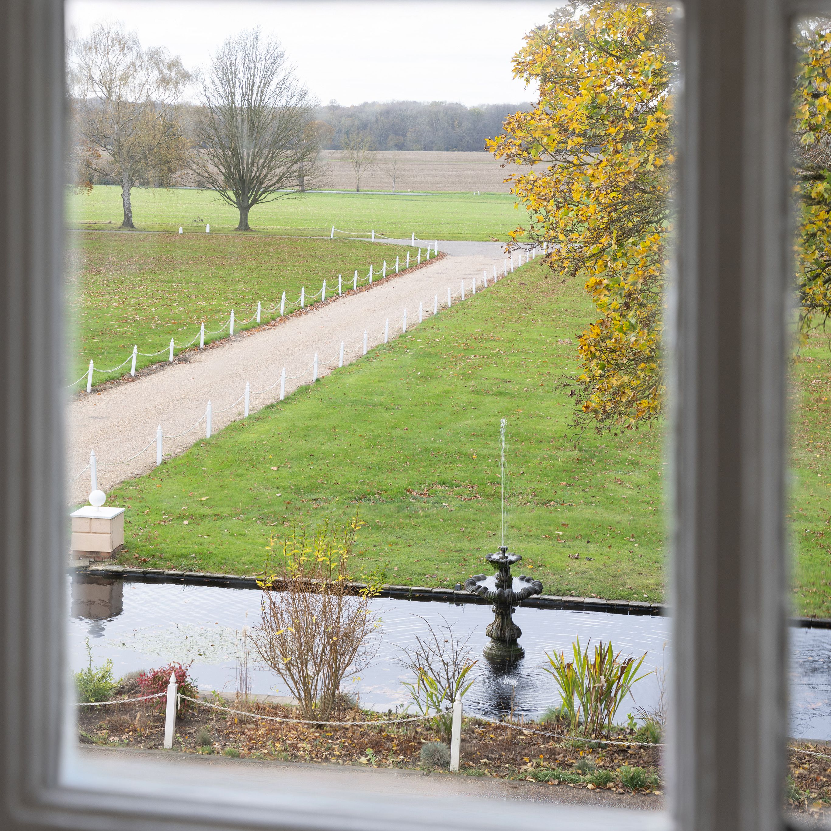 View through a window of a garden with a fountain and a long pathway flanked by grass and trees.