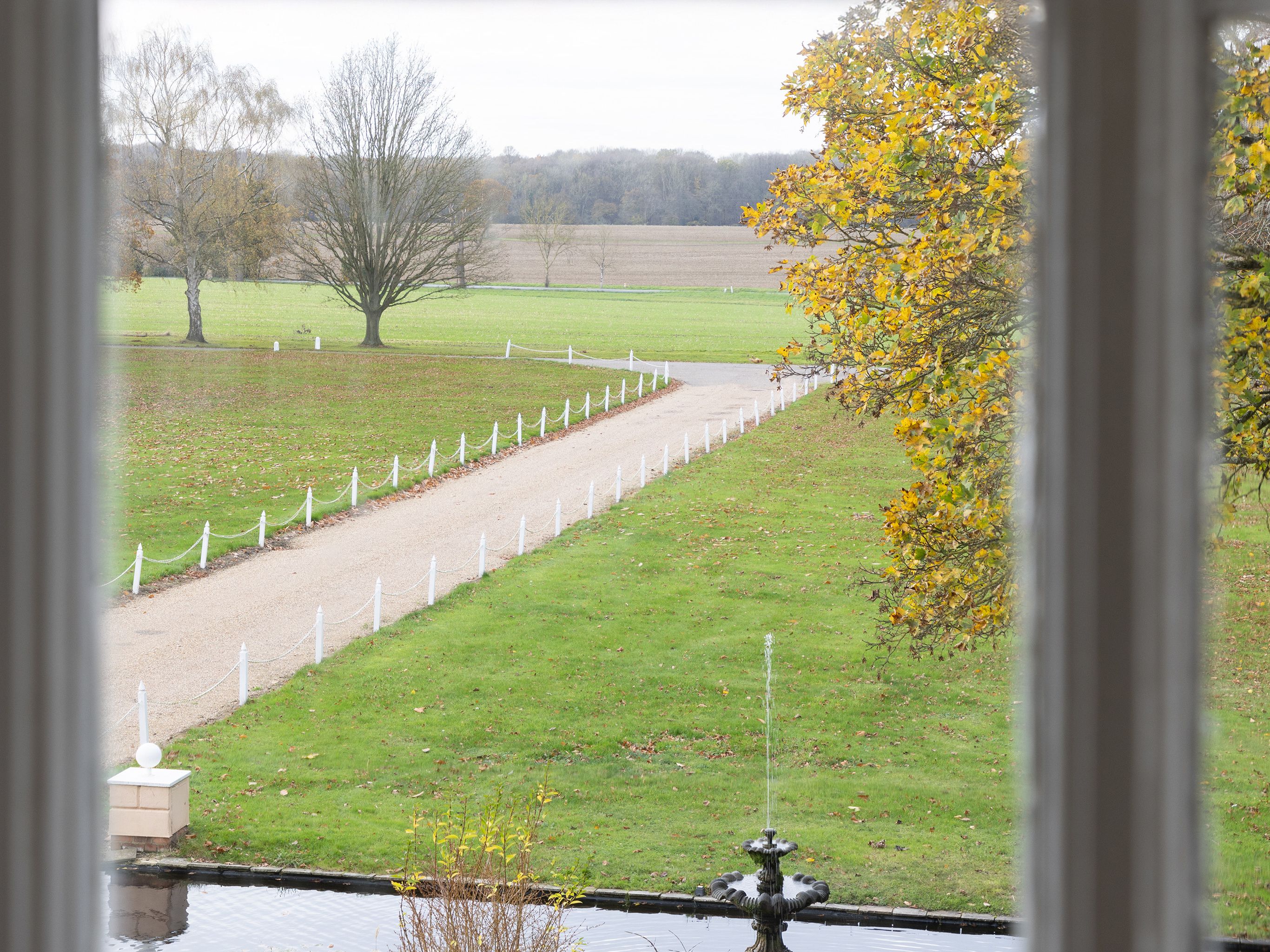 View through a window of a garden with a fountain and a long pathway flanked by grass and trees.