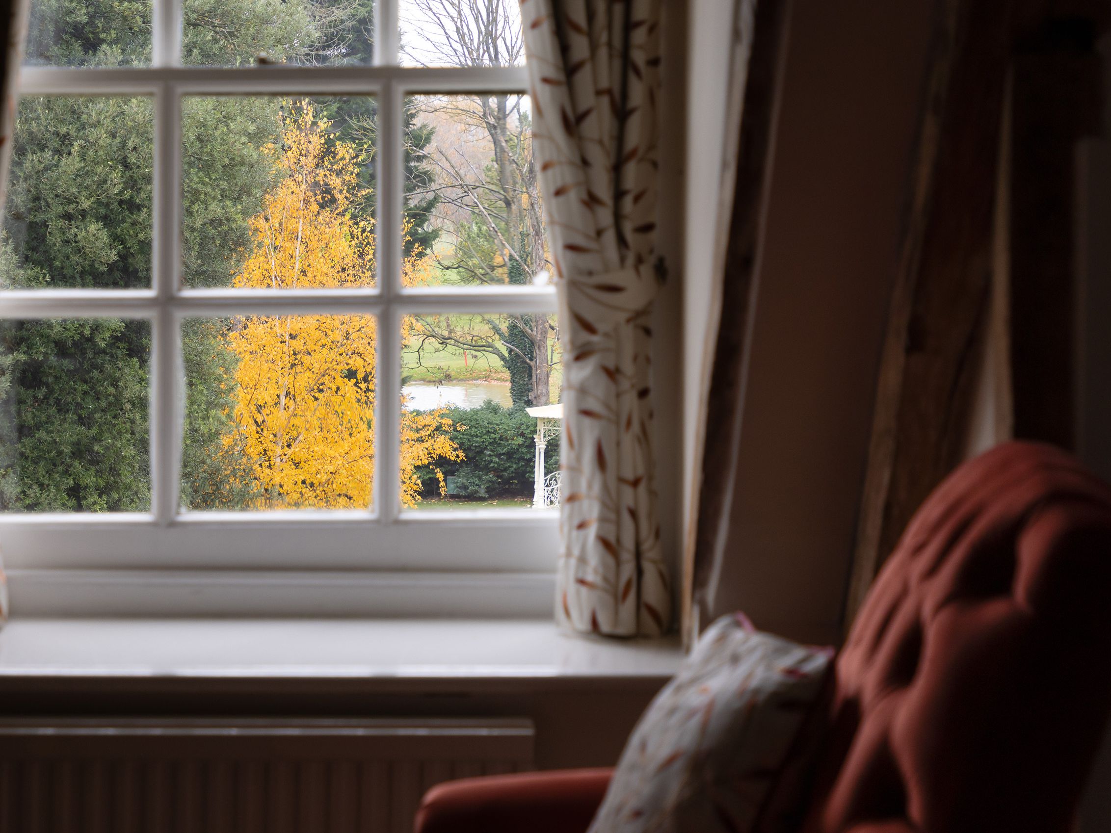 A cozy armchair by a window with curtains, overlooking a scenic garden with trees, including one with bright yellow autumn leaves.