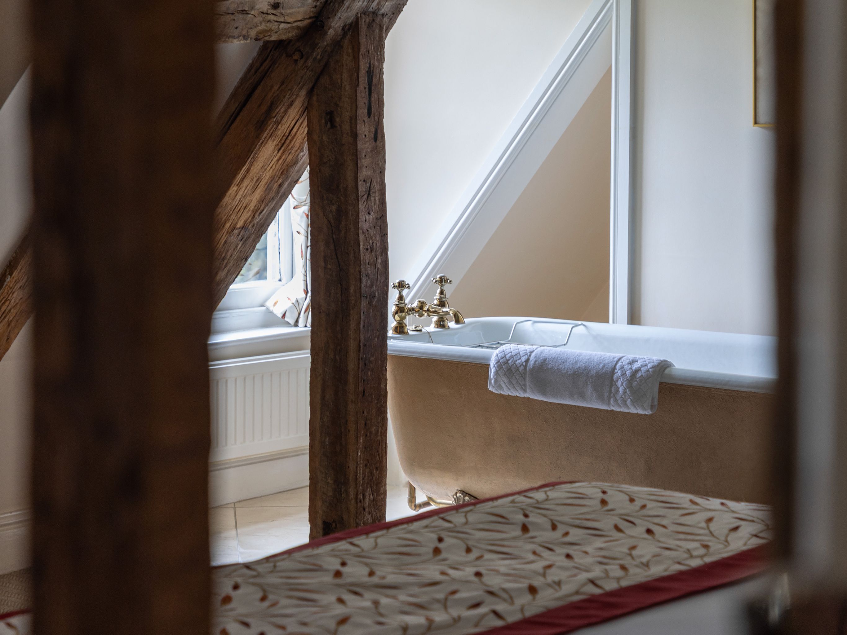 Rustic bathroom with exposed wooden beams and a freestanding bathtub under a slanted ceiling.