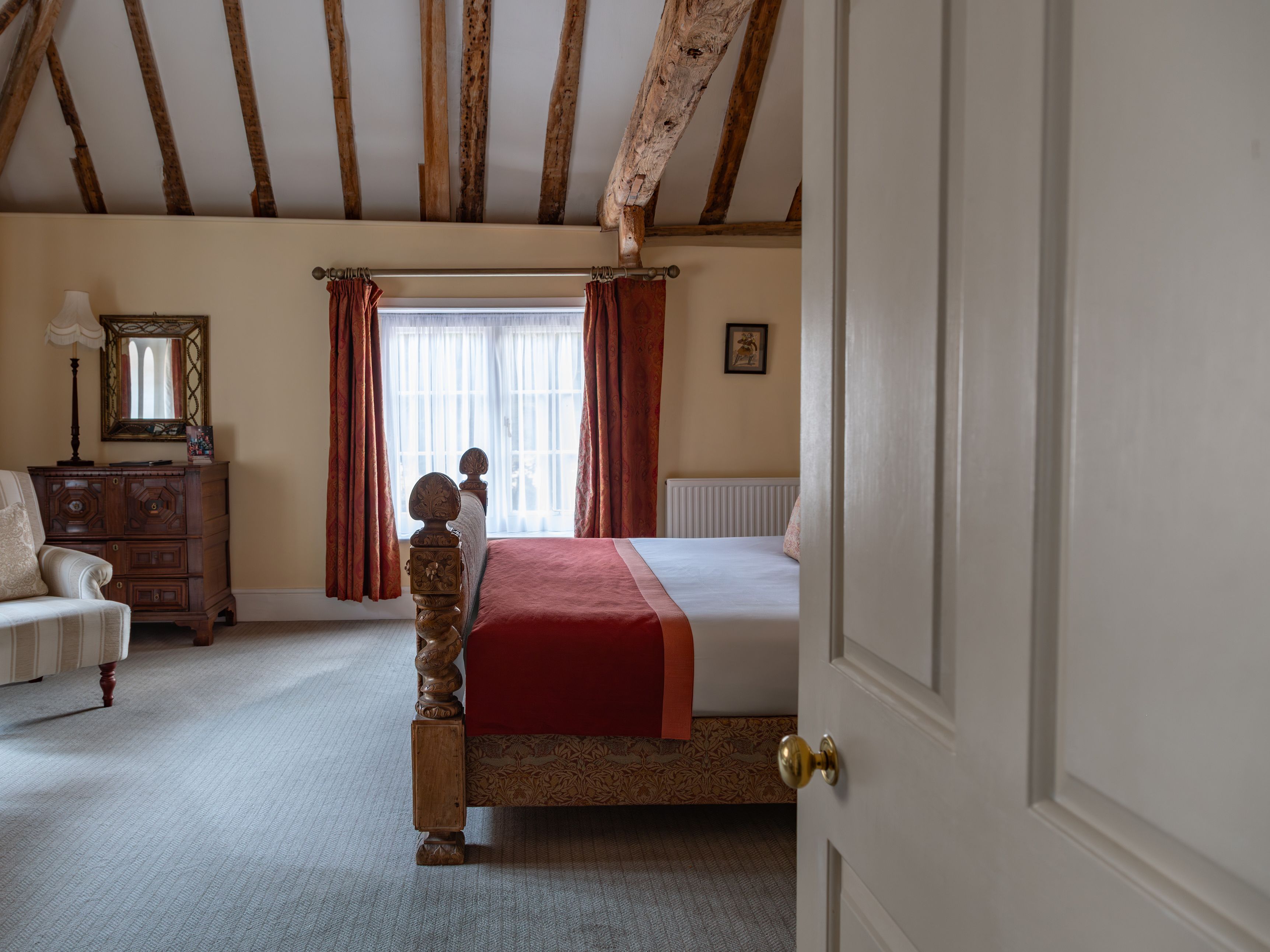 Cozy bedroom with exposed wooden beams, a large bed with a red blanket, antique furniture, and a window with red curtains.
