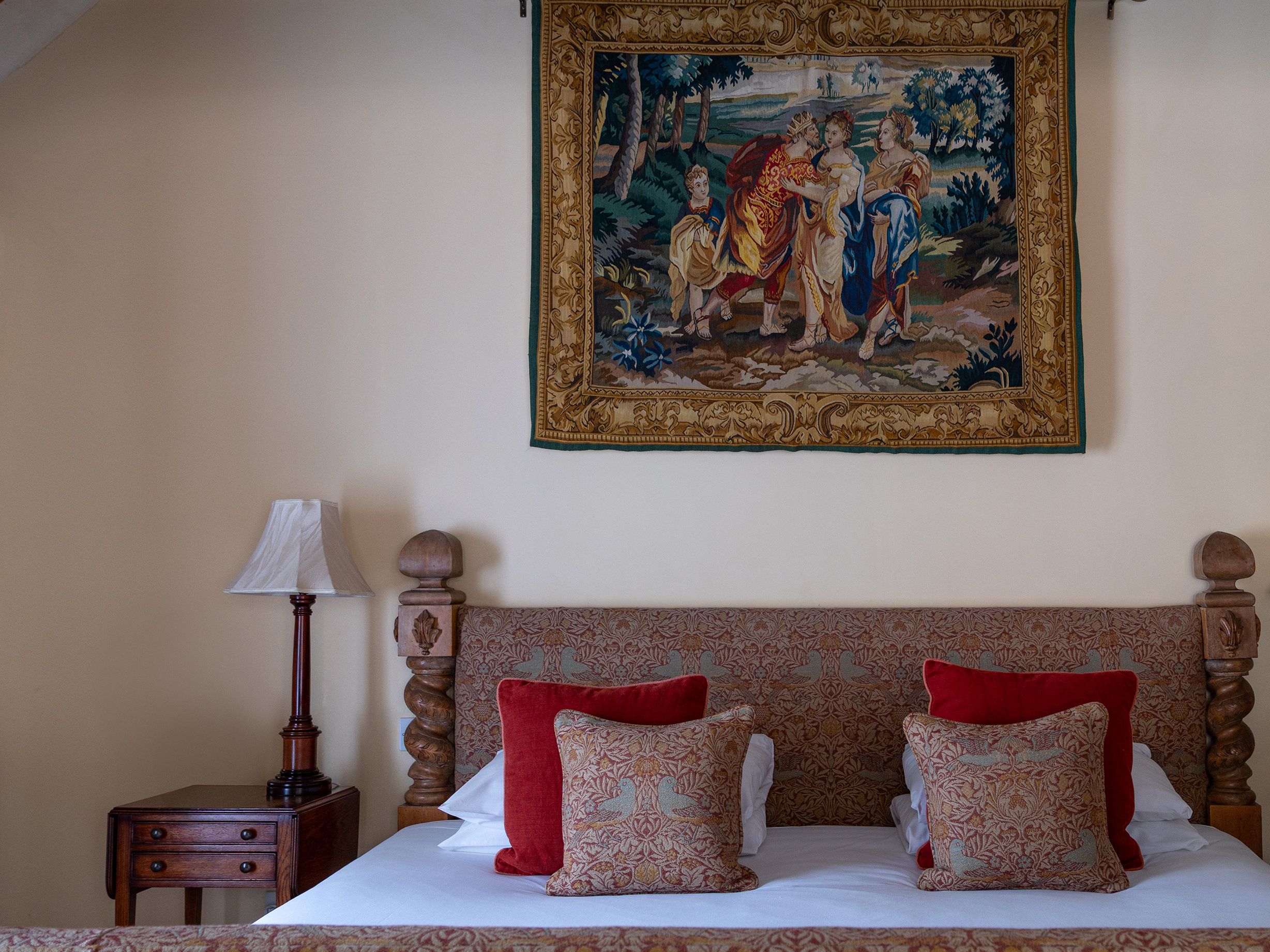 Cozy bedroom with a patterned bed, red and gold pillows, an antique wooden nightstand, and a decorative wall tapestry above the bed.