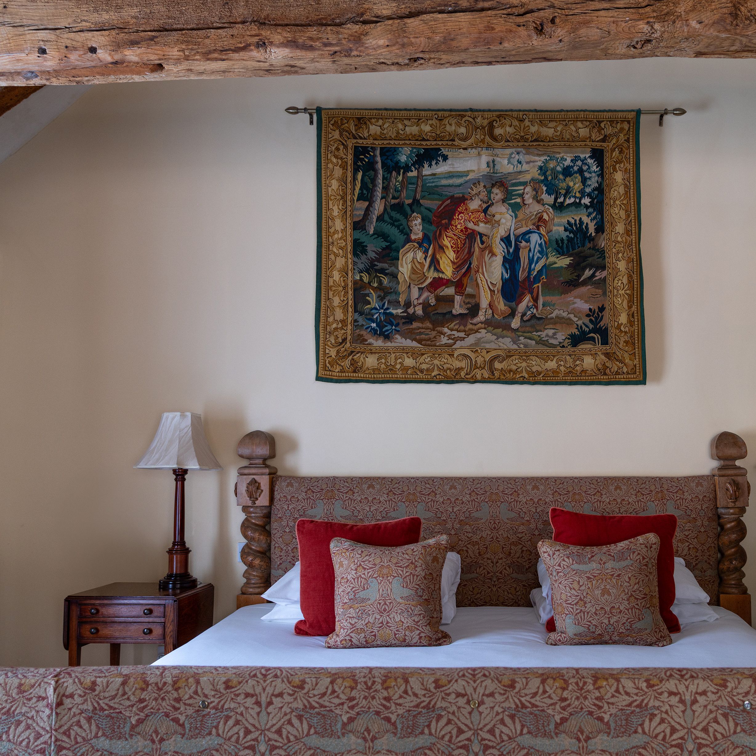 Cozy bedroom with a patterned bed, red and gold pillows, an antique wooden nightstand, and a decorative wall tapestry above the bed.