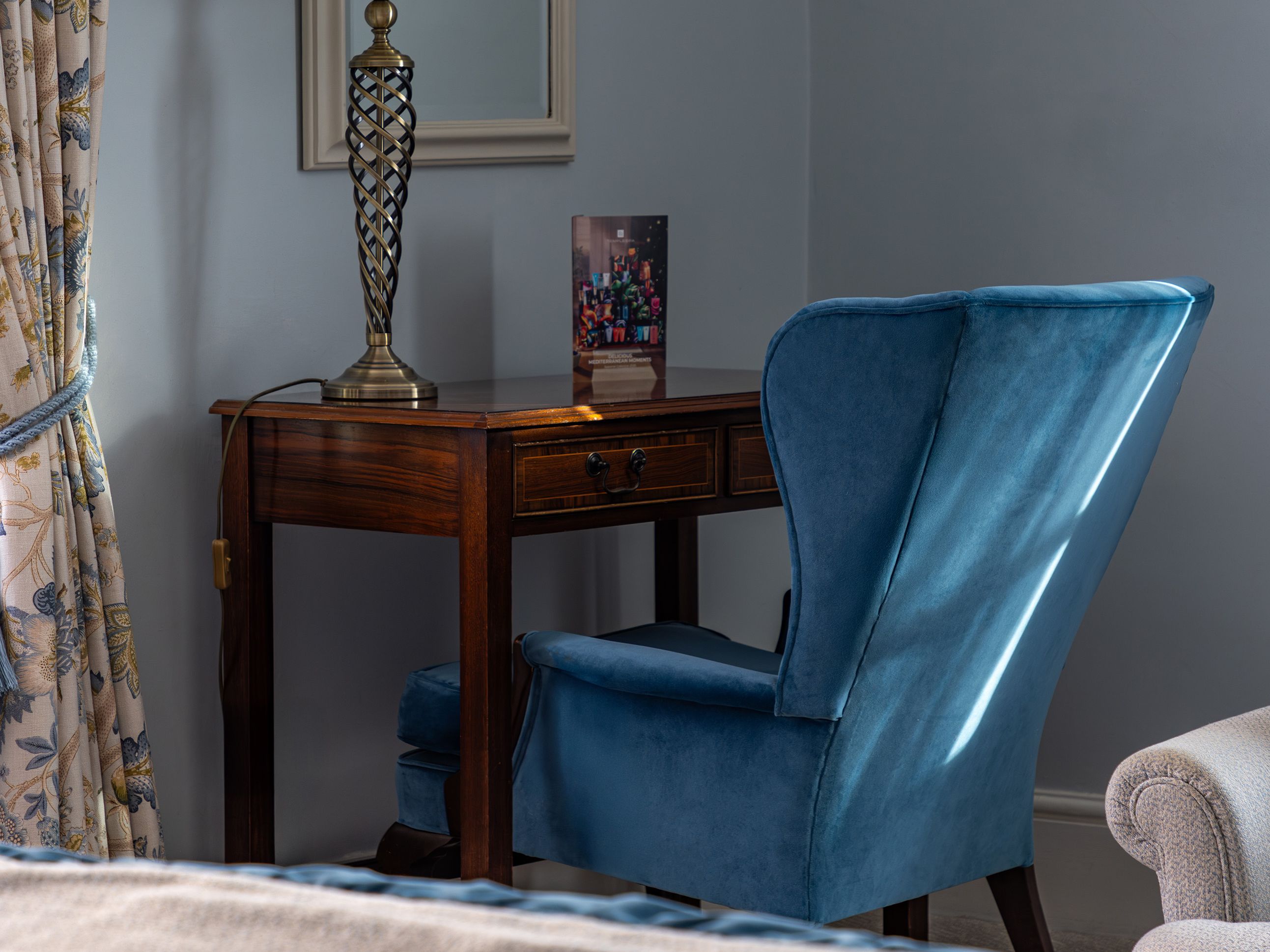 A cozy bedroom corner with a blue velvet chair, wooden desk, lamp, mirror, and floral curtains.