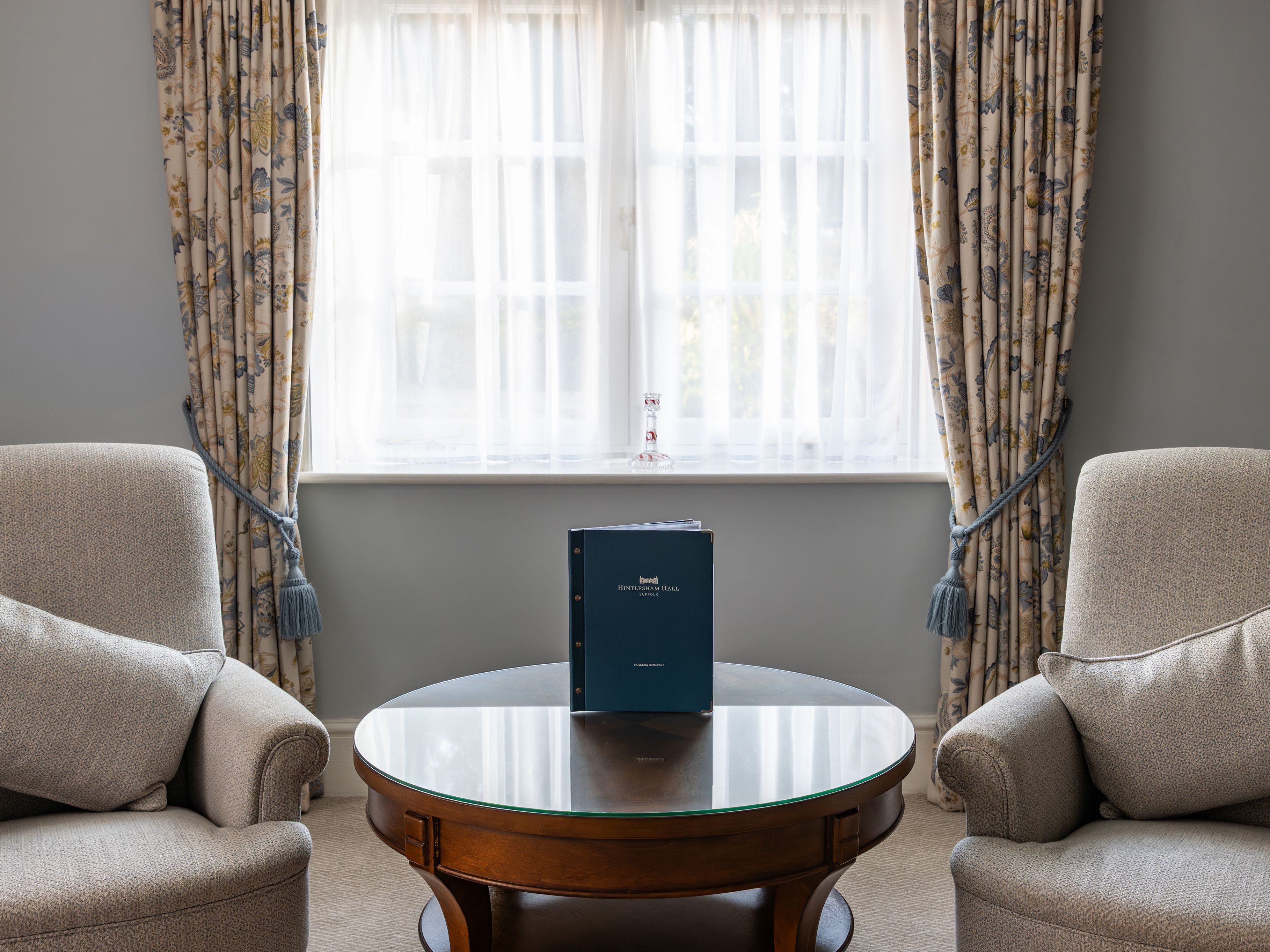Elegant sitting area with two armchairs, a round glass coffee table, and a binder placed in front of a window with floral curtains.