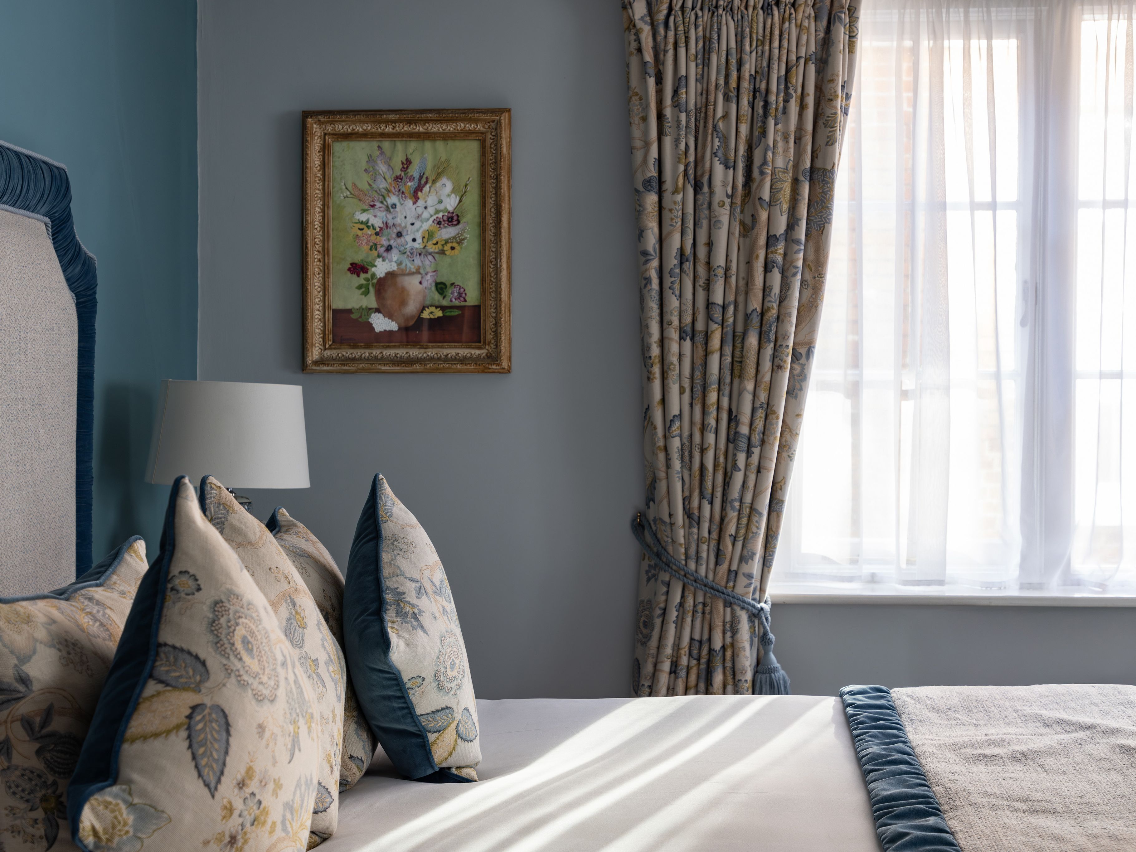 A cozy bedroom with a neatly made bed, floral cushions, a floral painting on the wall, and patterned curtains next to a sunlit window.