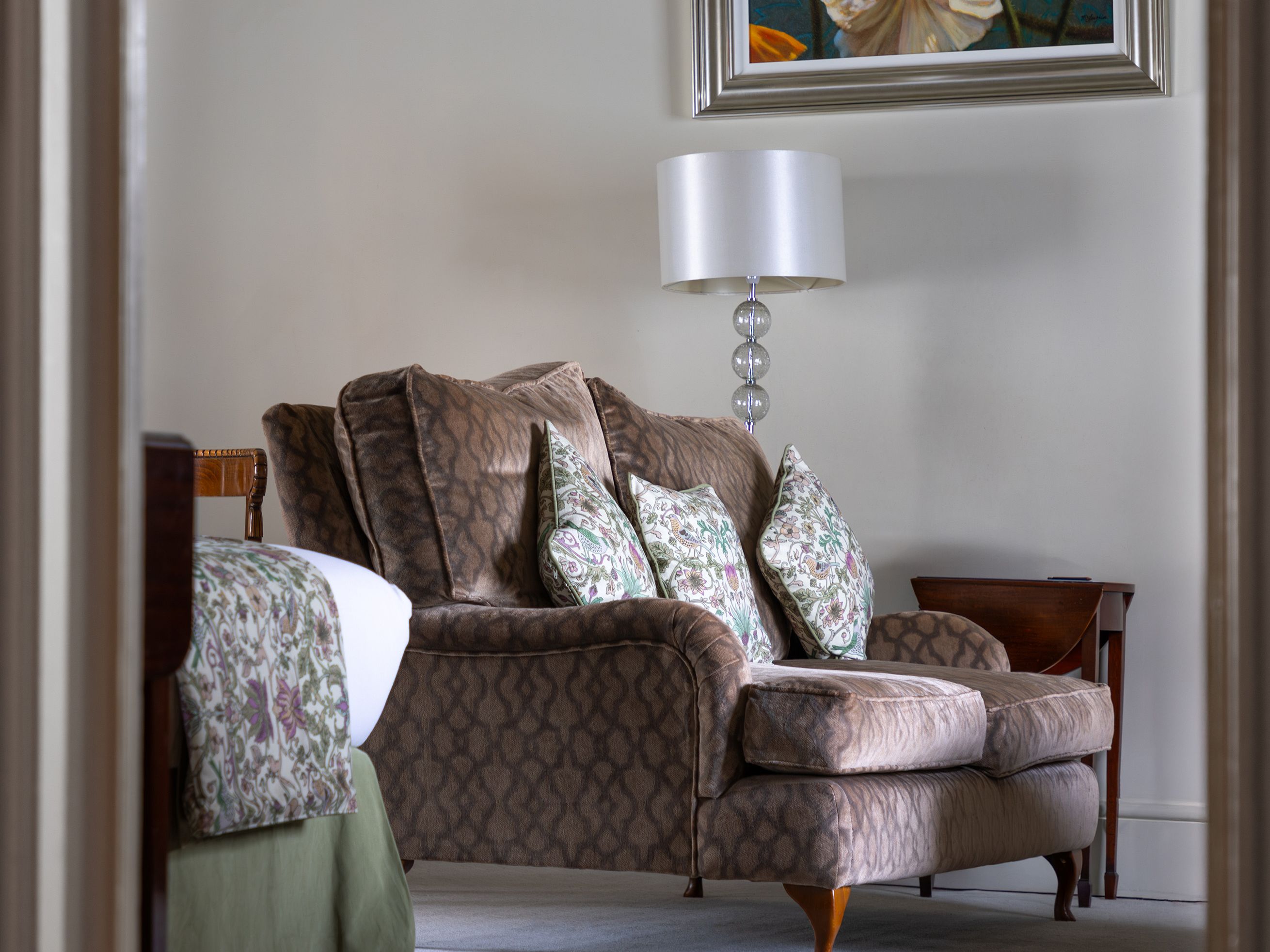 Elegant living room with patterned couch, floral pillows, white lamp and a framed flower painting.