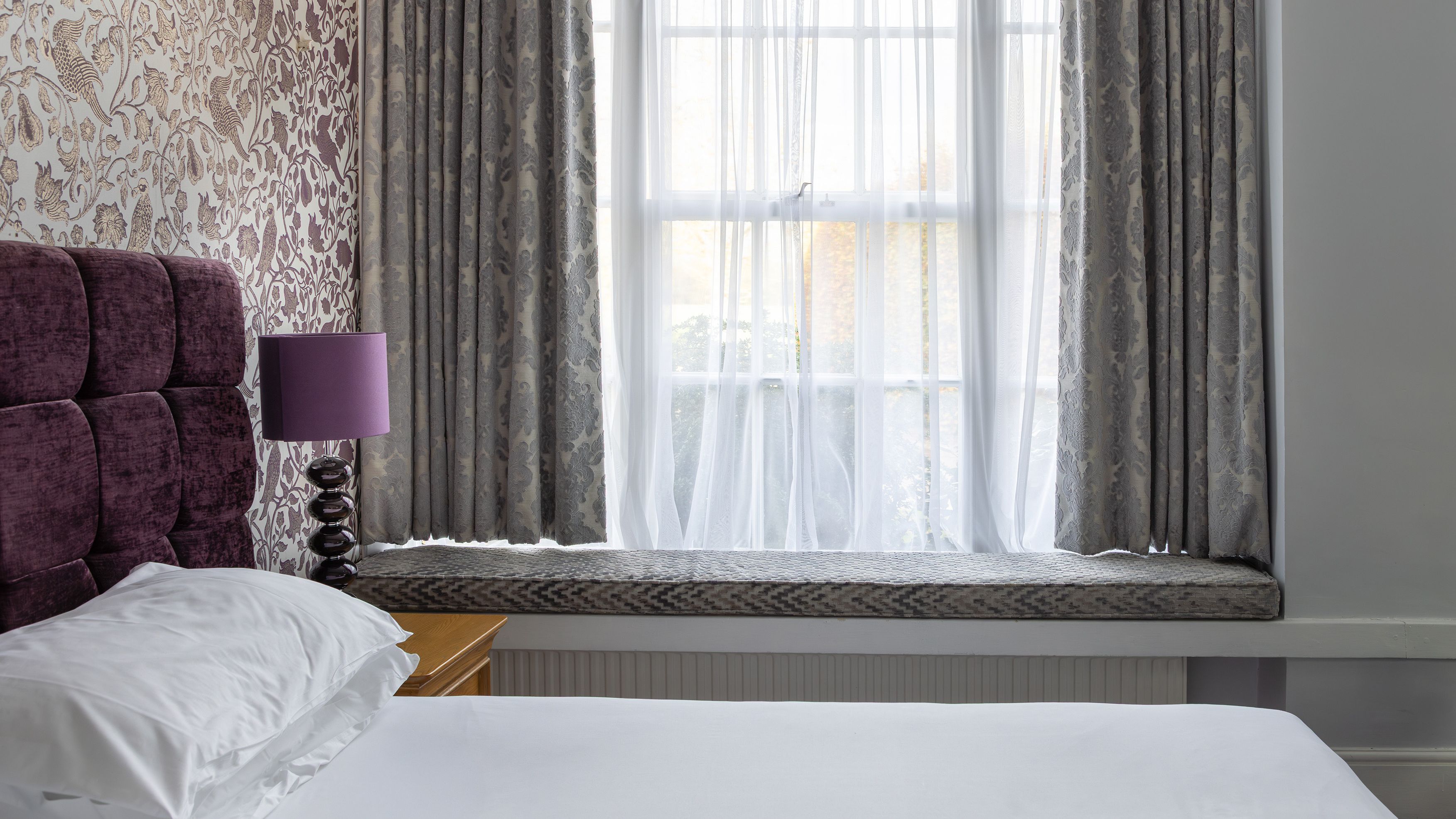 Modern bedroom with purple headboard, patterned wallpaper, large window with curtains, window seat, and purple lamp.