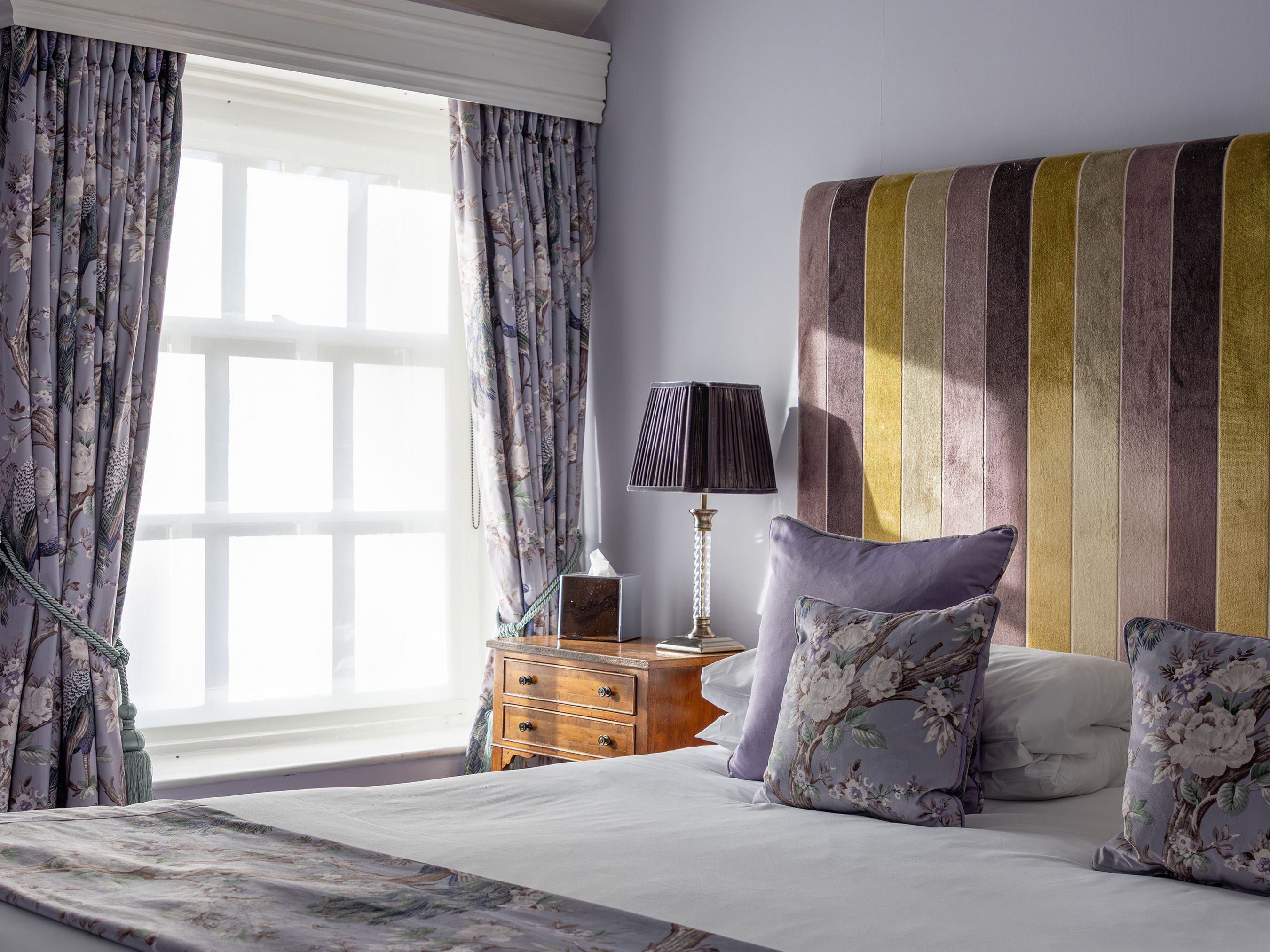Elegant bedroom with purple walls, floral curtains, and a striped upholstered headboard.