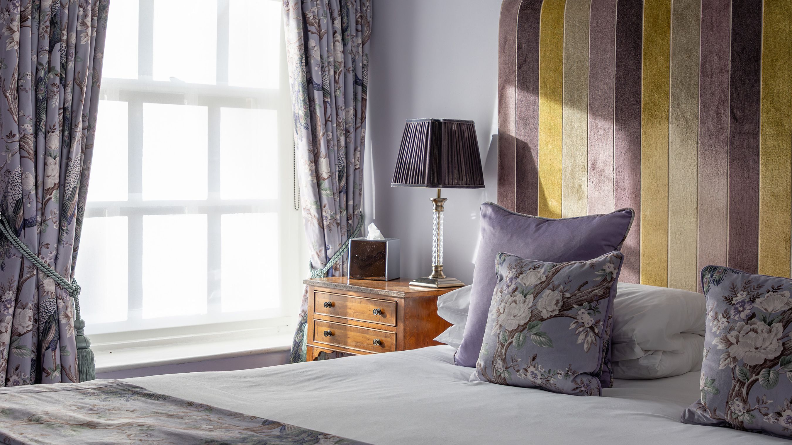 Elegant bedroom with purple walls, floral curtains, and a striped upholstered headboard.
