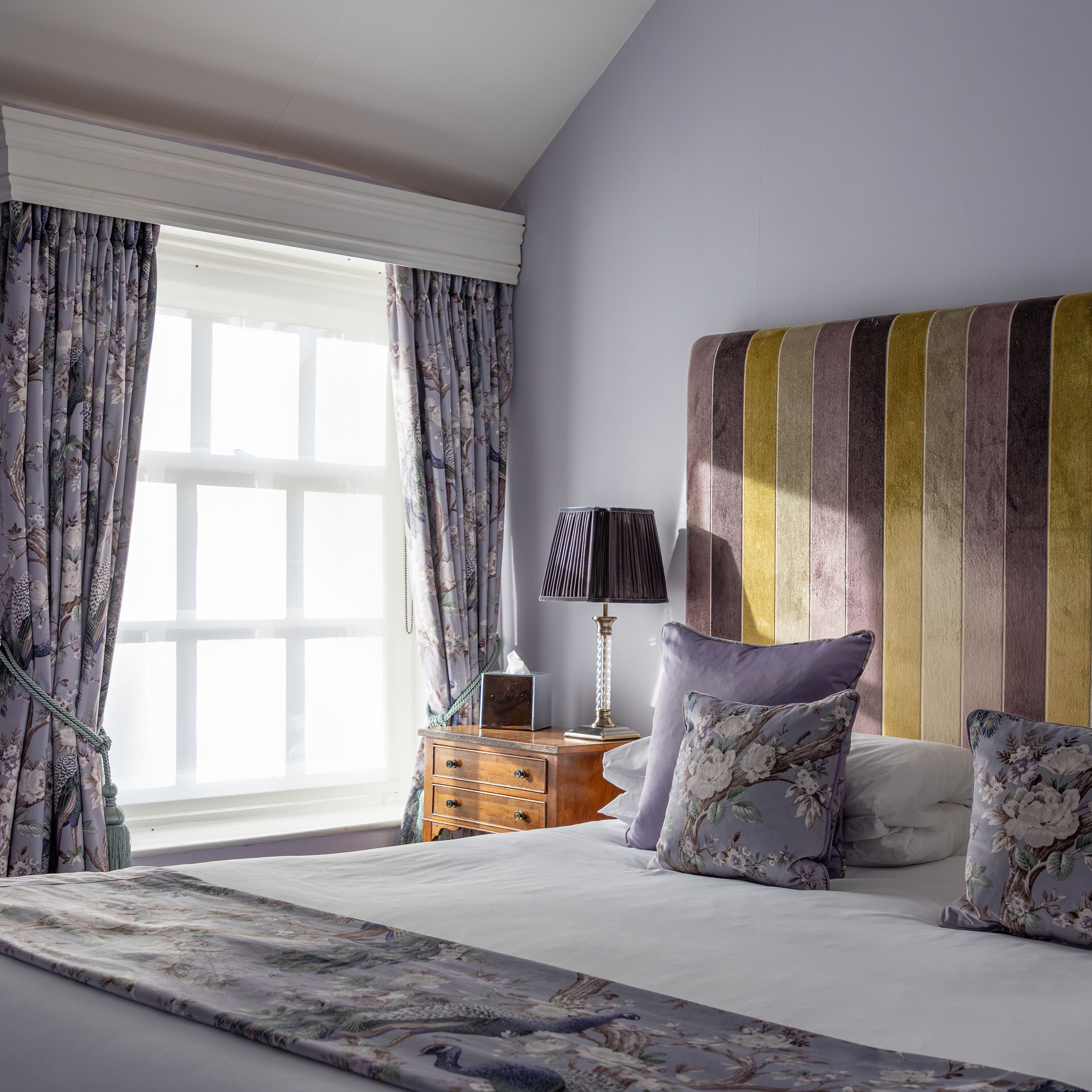 Elegant bedroom with purple walls, floral curtains, and a striped upholstered headboard.