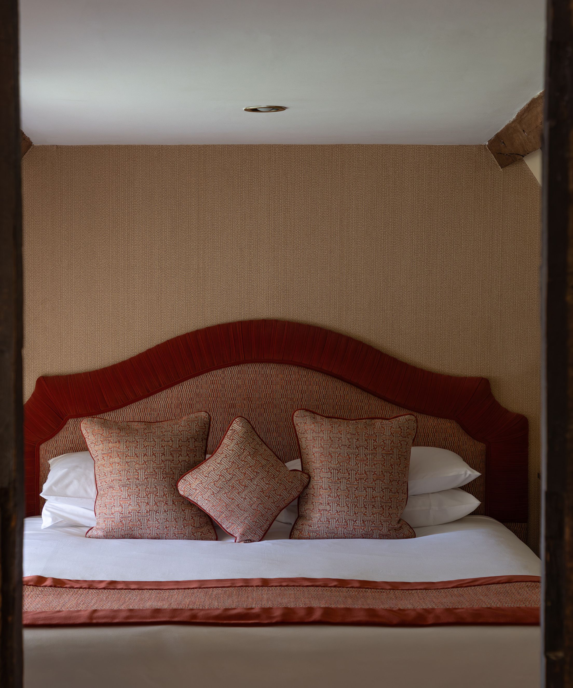 Cozy attic bedroom with wooden beams, a bed with red headboard and cushions, and bedside tables.