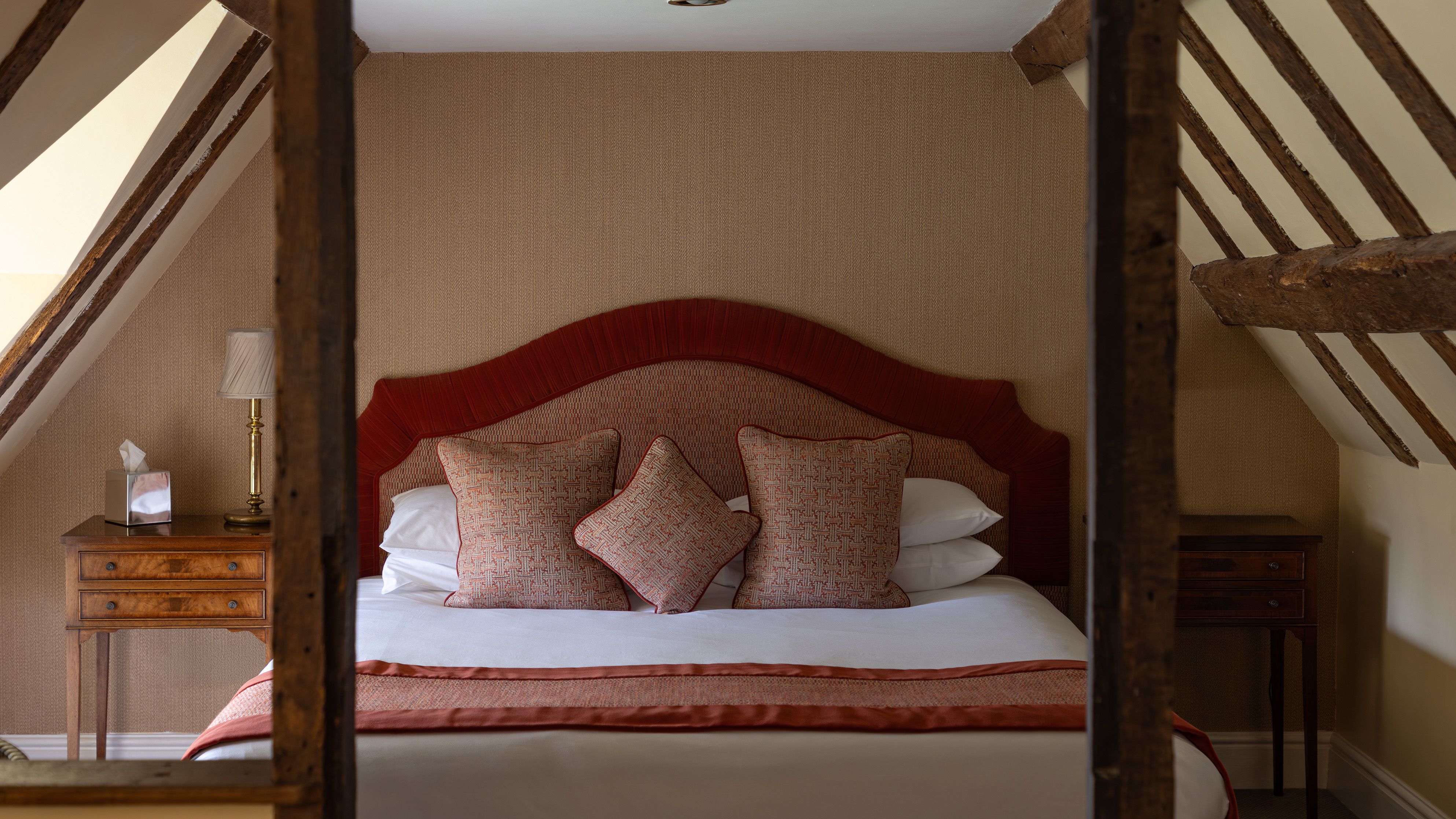 Cozy attic bedroom with wooden beams, a bed with red headboard and cushions, and bedside tables.