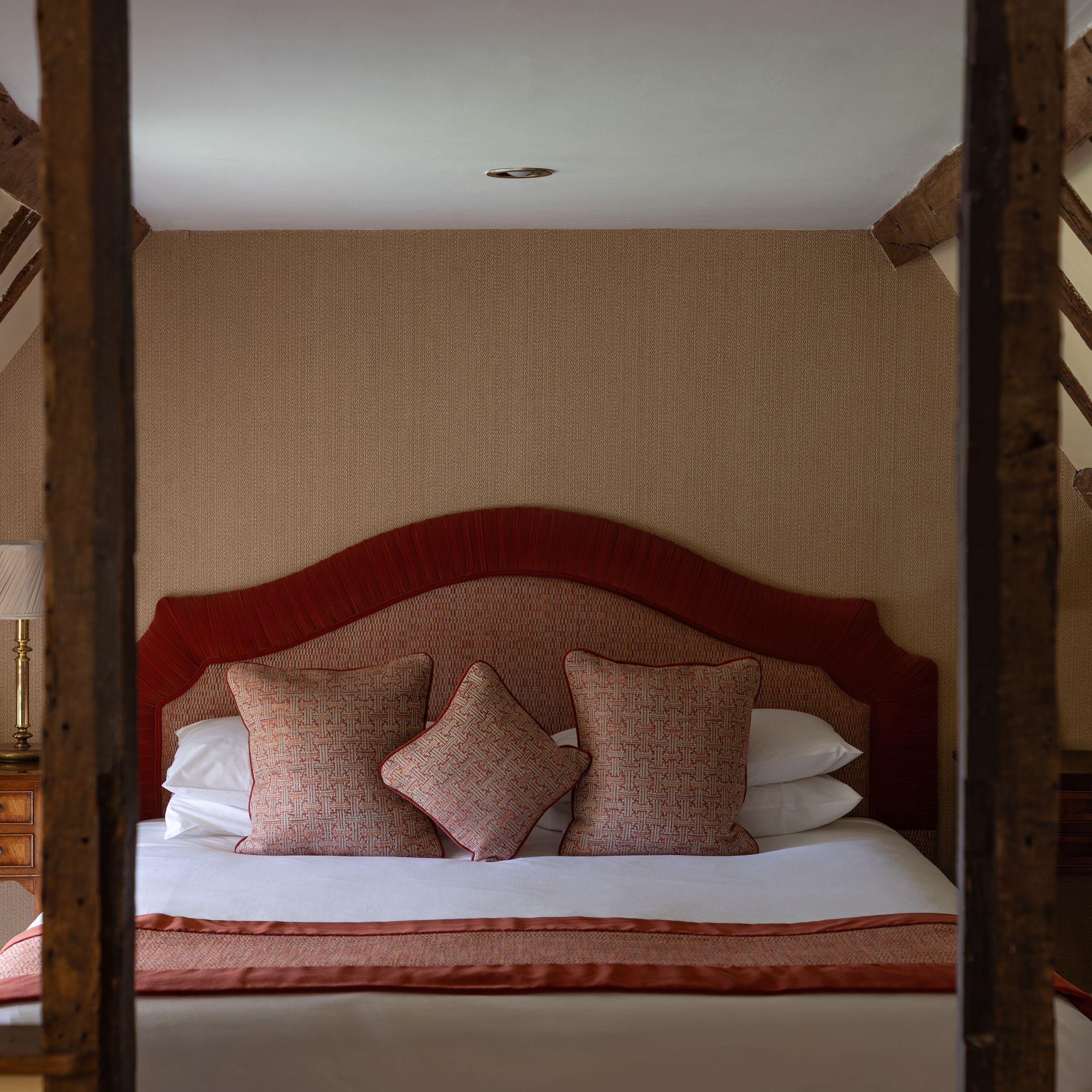 Cozy attic bedroom with wooden beams, a bed with red headboard and cushions, and bedside tables.