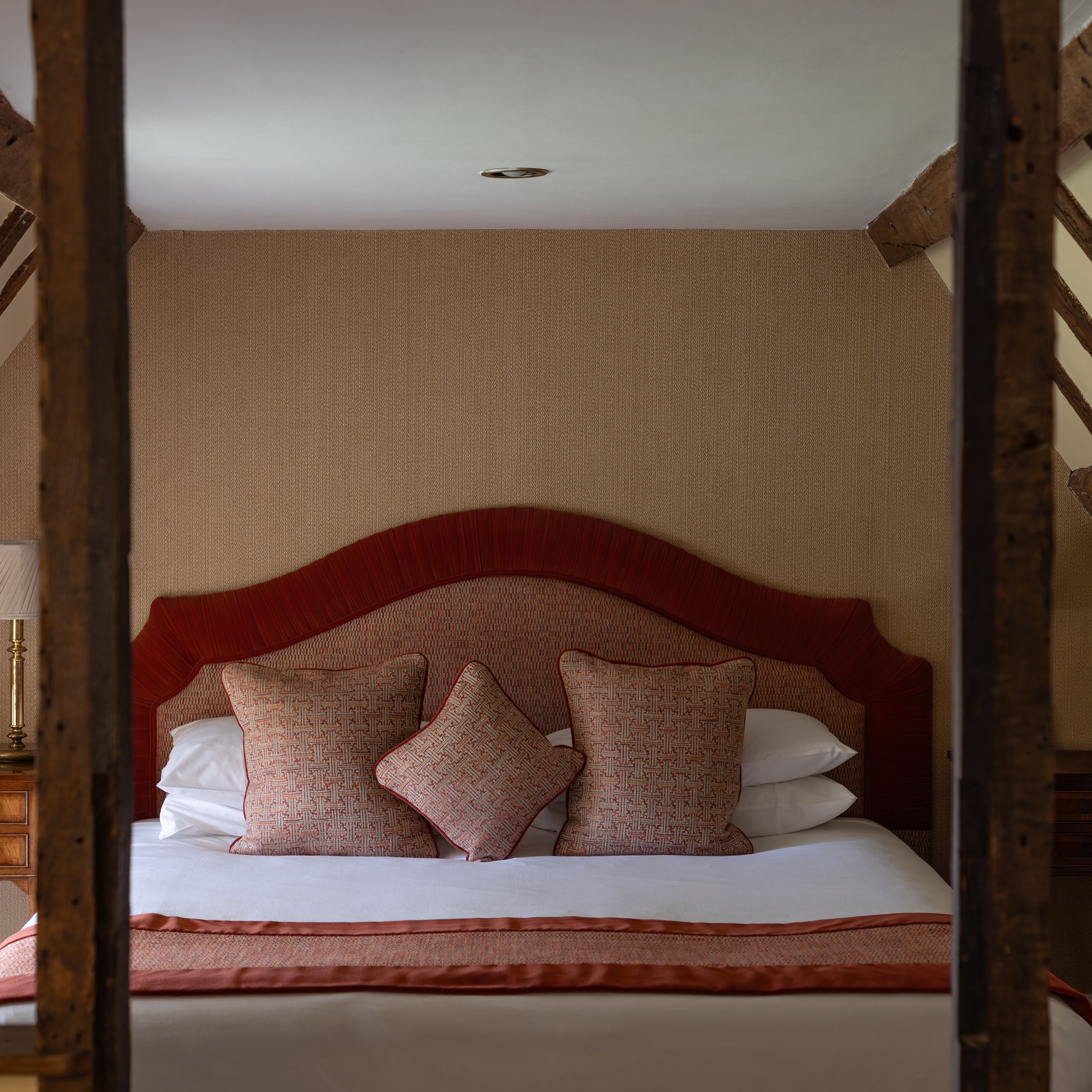 Cozy attic bedroom with wooden beams, a bed with red headboard and cushions, and bedside tables.